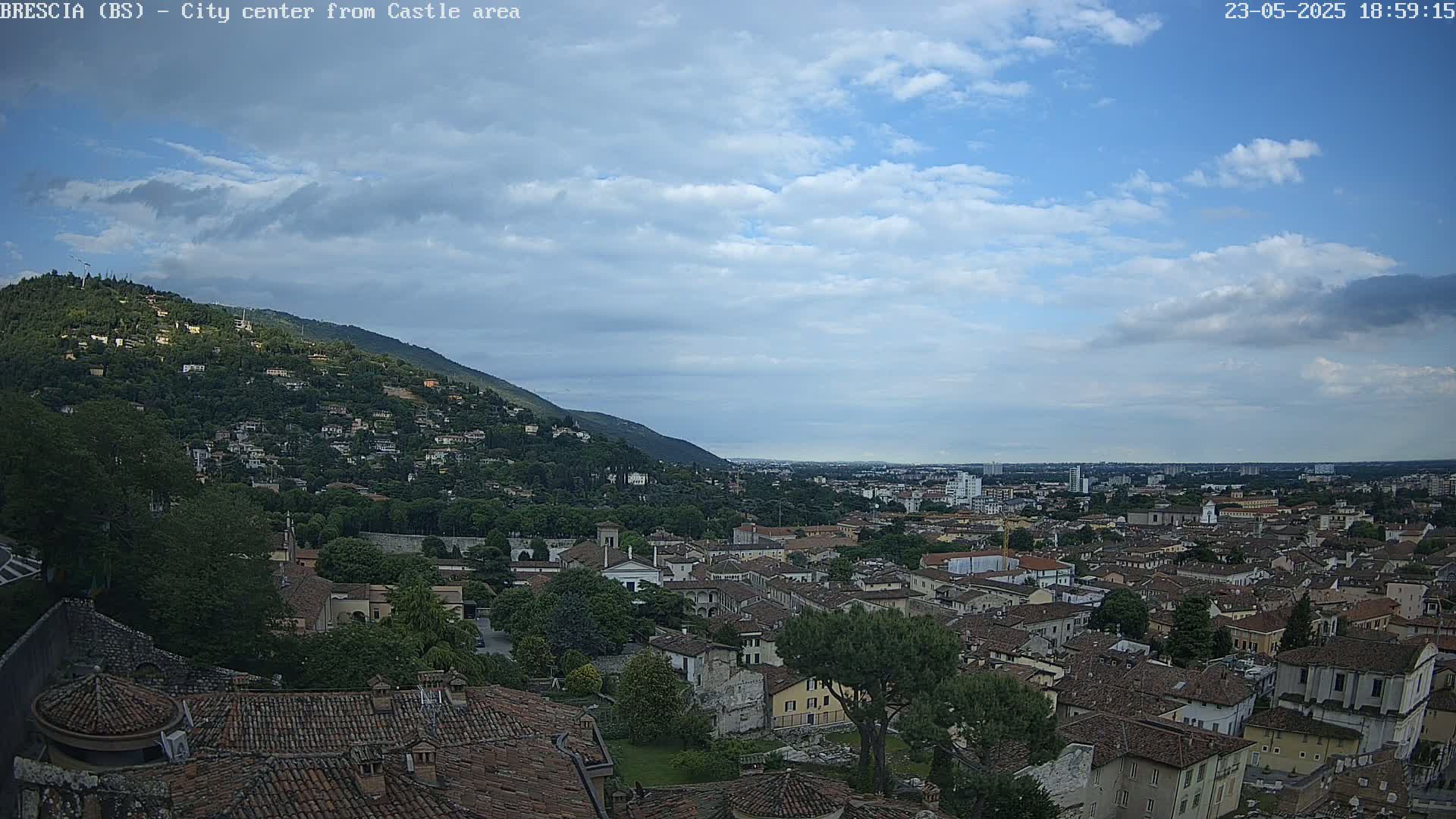 A partly cloudy sky overlooks a sprawling city nestled against a hillside, with many brown-roofed buildings visible.