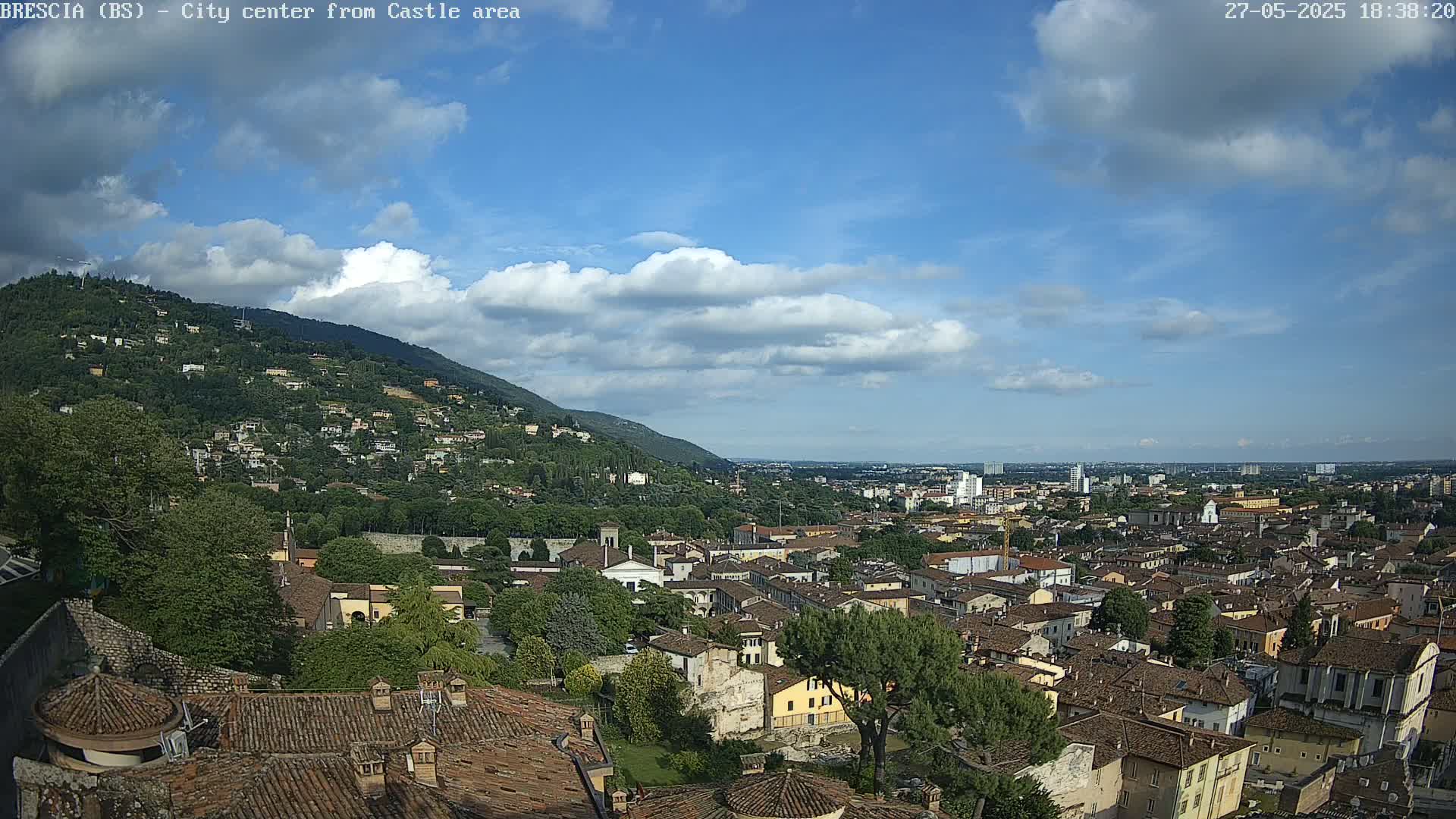 A sprawling city landscape, nestled against a verdant hill under a partly cloudy blue sky.