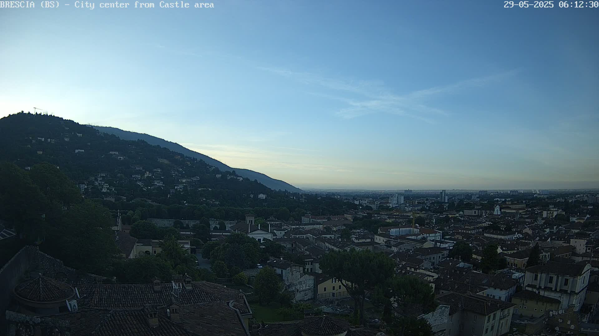 A city sprawls beneath a clear, blue sky at dawn, nestled against a hill covered in trees and buildings.