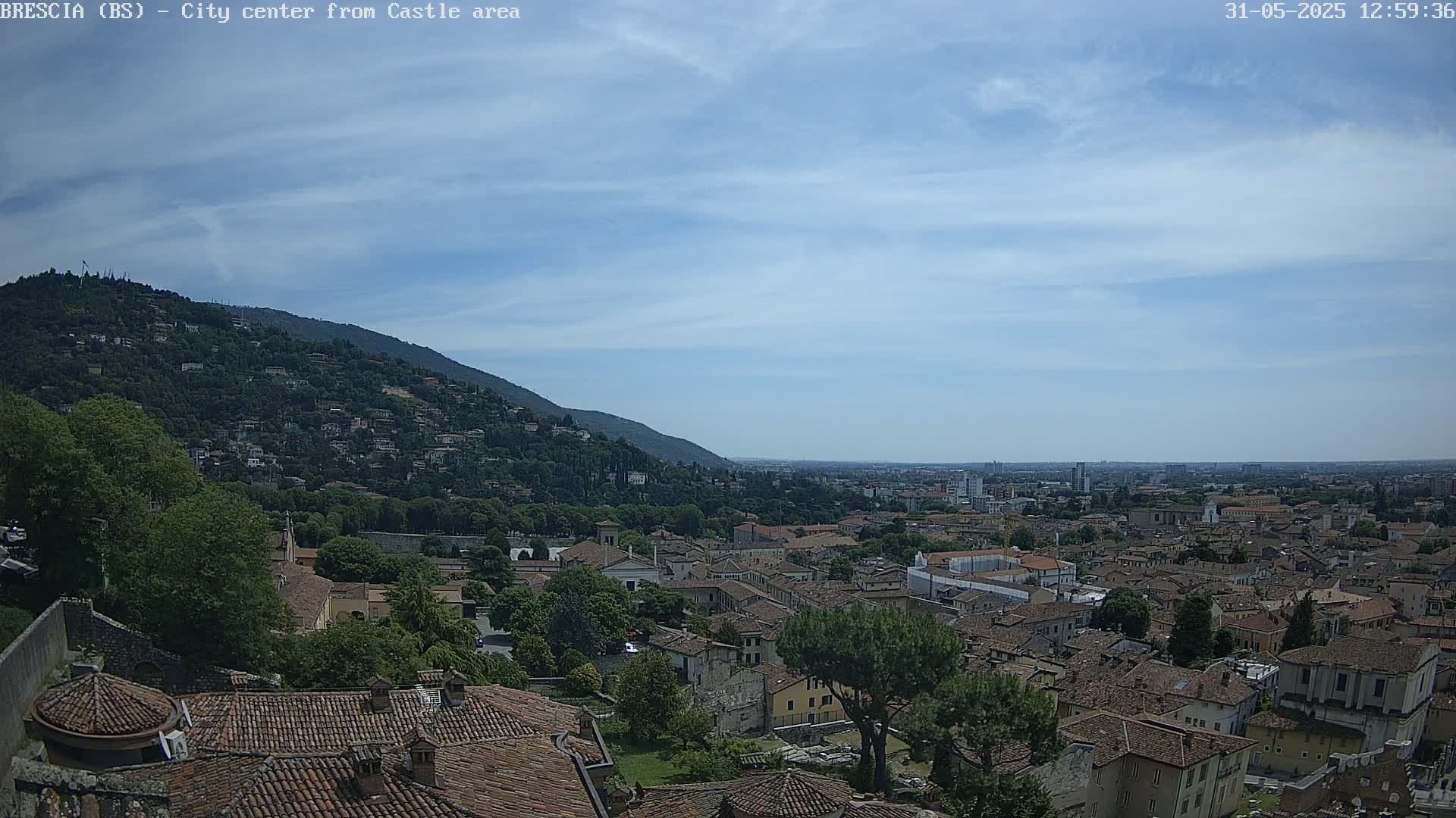 A sprawling city with terracotta rooftops nestles beneath a hill covered in lush vegetation under a mostly clear, light blue sky.