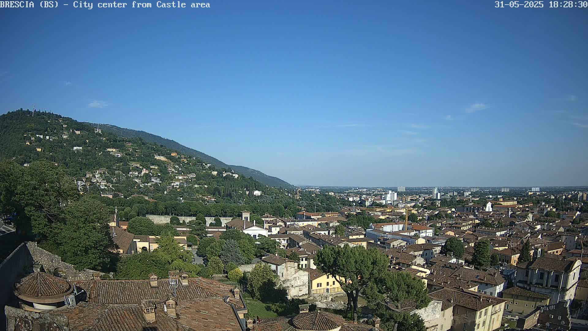 A sprawling city with red-tiled rooftops nestled beneath a clear blue sky and a verdant hill.