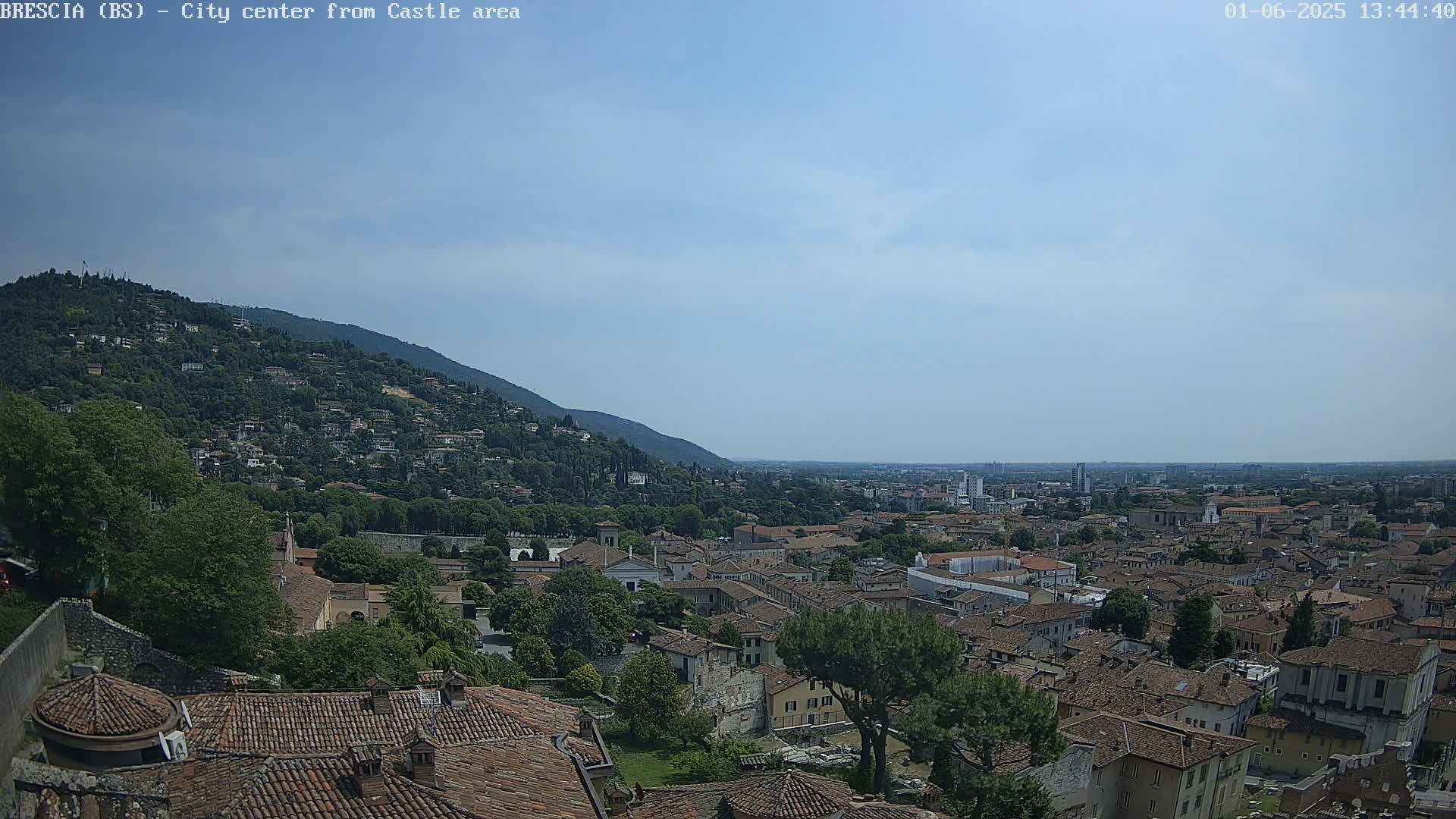 A sprawling city with red-tiled roofs nestles at the base of a green hill under a mostly clear, light blue sky.