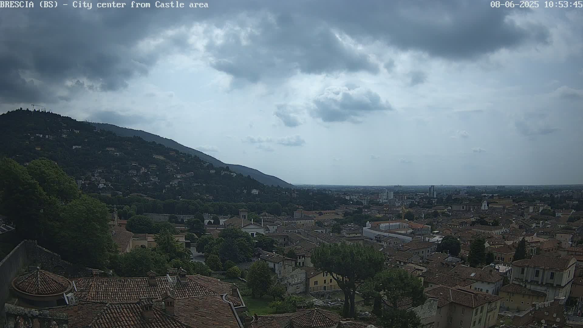 A sprawling city with red-tiled roofs nestled against a green hill under a mostly cloudy sky.