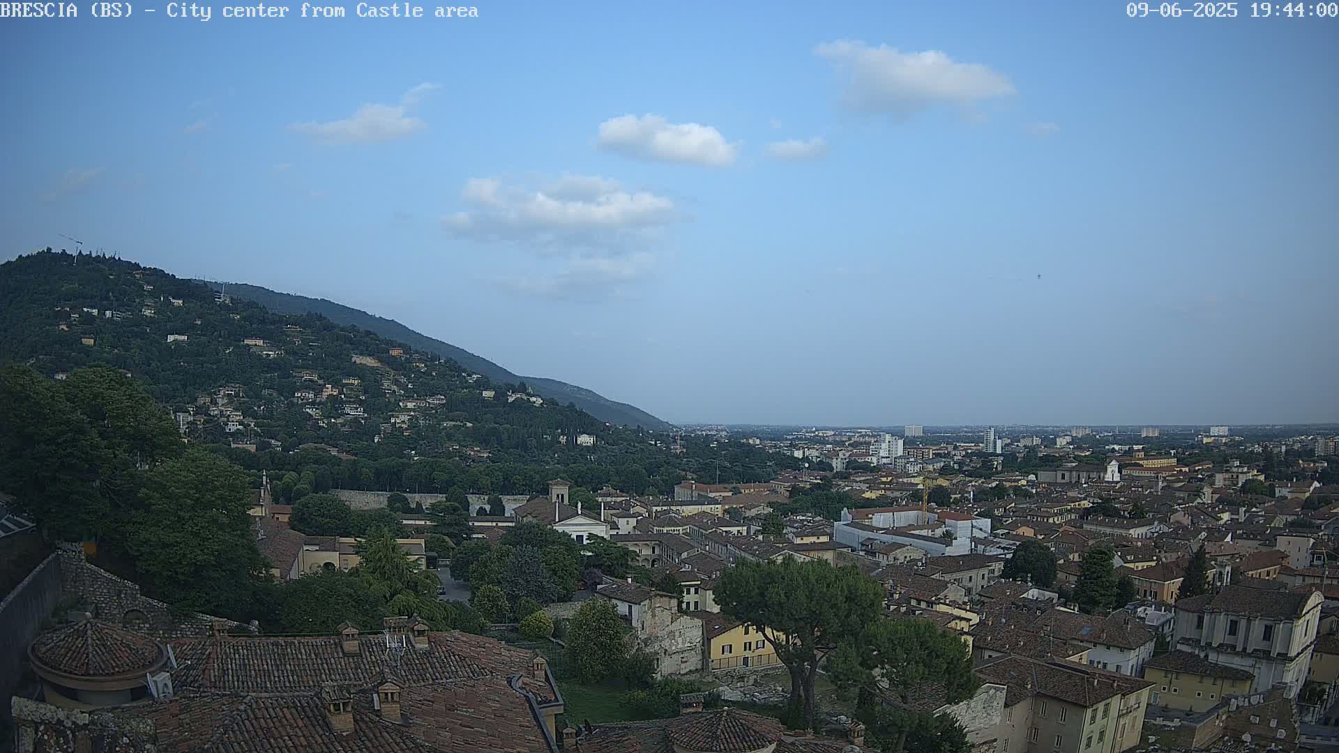 A sprawling city with terracotta rooftops nestles beneath a hill covered in trees and houses, all under a mostly clear, light-blue sky with a few scattered clouds.