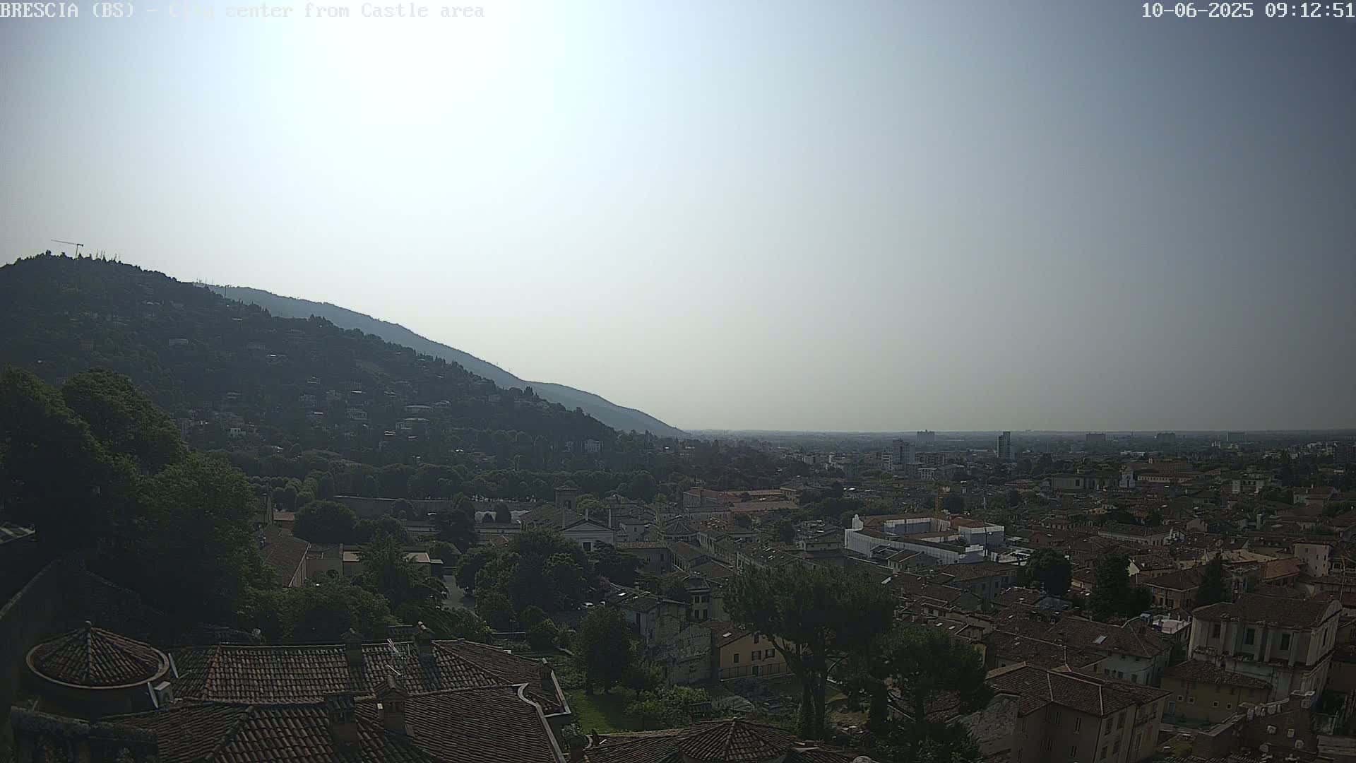 A hazy, sunny day overlooks a sprawling city nestled at the foot of a large, tree-covered hill.