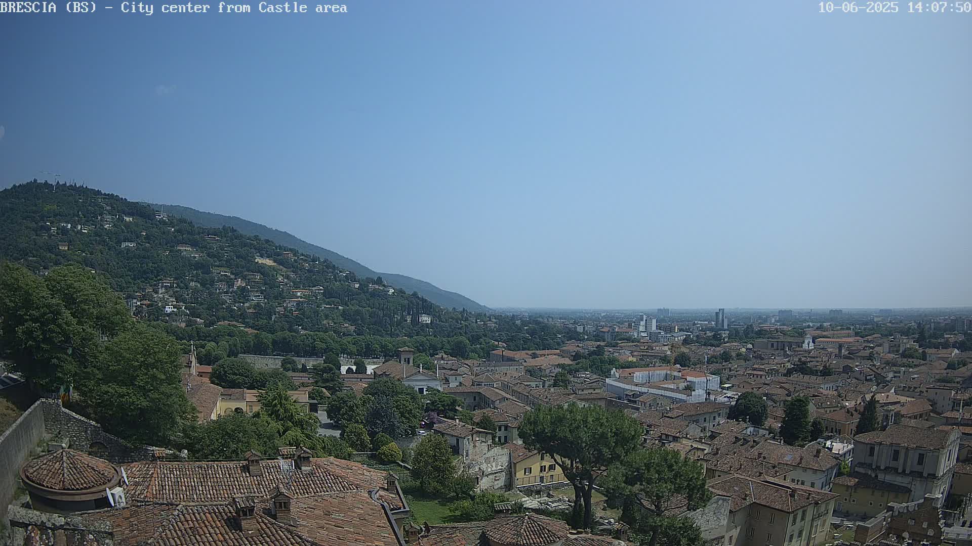 A sprawling city with red-tiled roofs nestled against a verdant hillside under a clear blue sky.