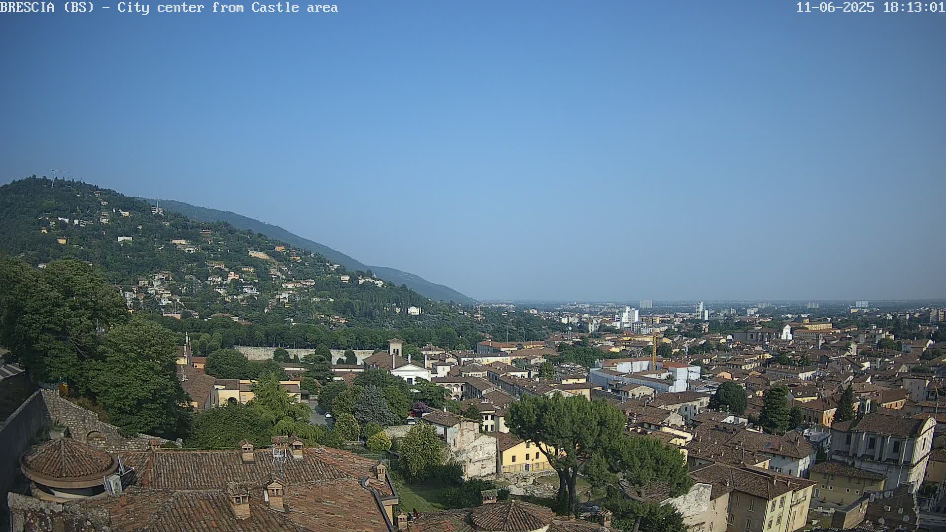 A sprawling city scene under a clear blue sky, viewed from a hilltop vantage point, shows a densely packed cityscape with red-tiled roofs, interspersed with green trees and a distant mountain range.
