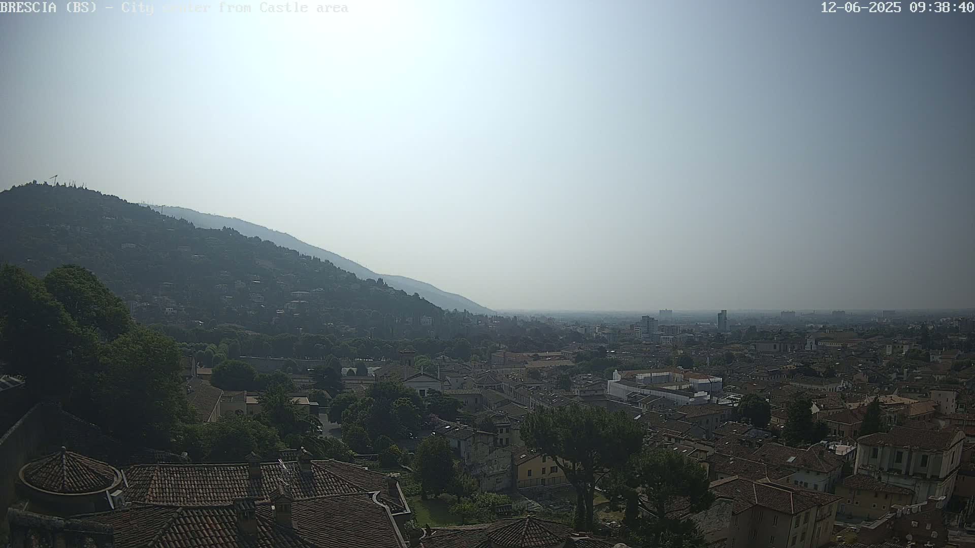 A hazy, sunny view shows a sprawling city nestled at the base of a large hill, with rooftops and trees dominating the foreground.