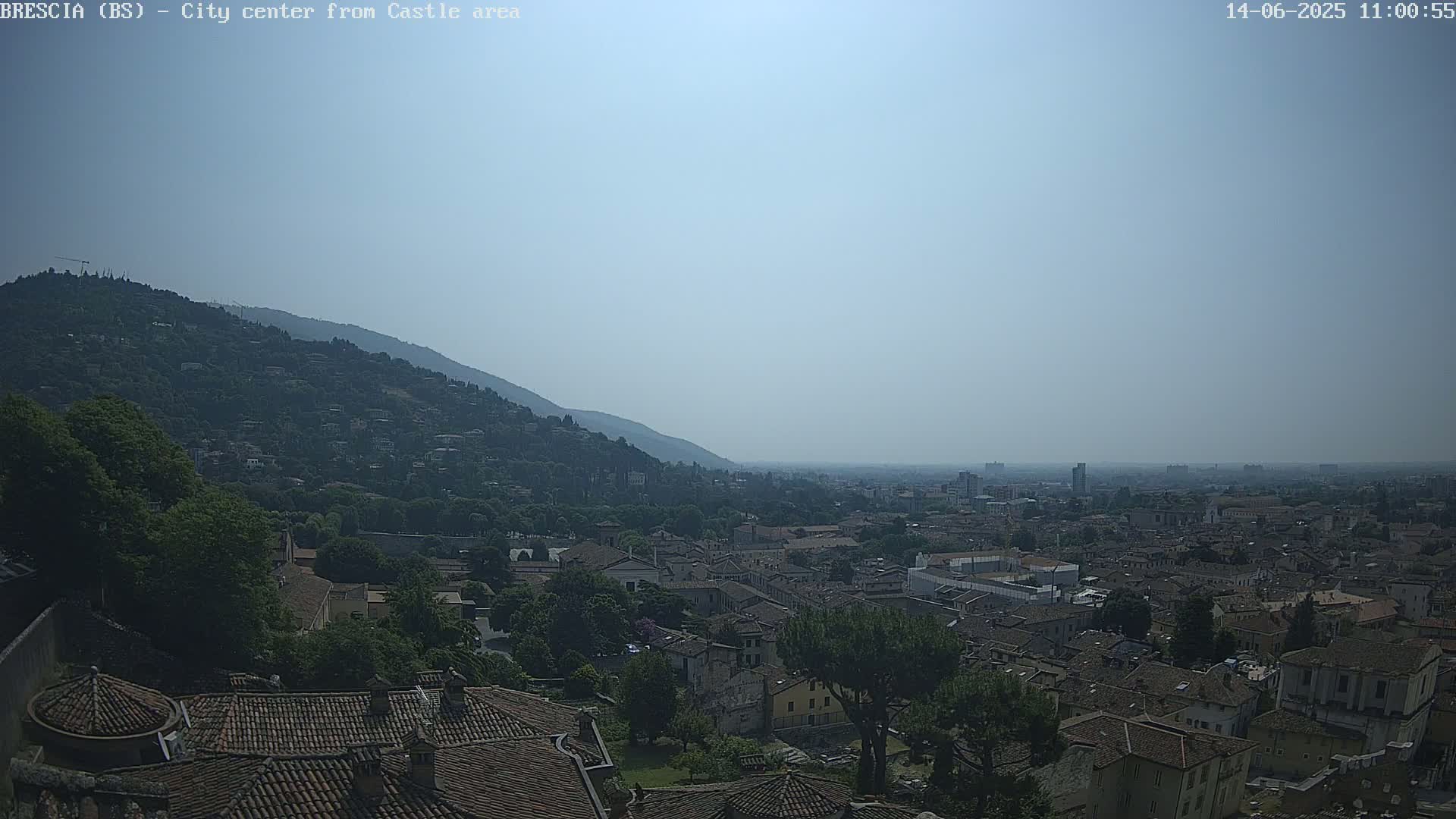 A hazy, sunny day reveals a sprawling city nestled below a verdant hill, viewed from an elevated vantage point.