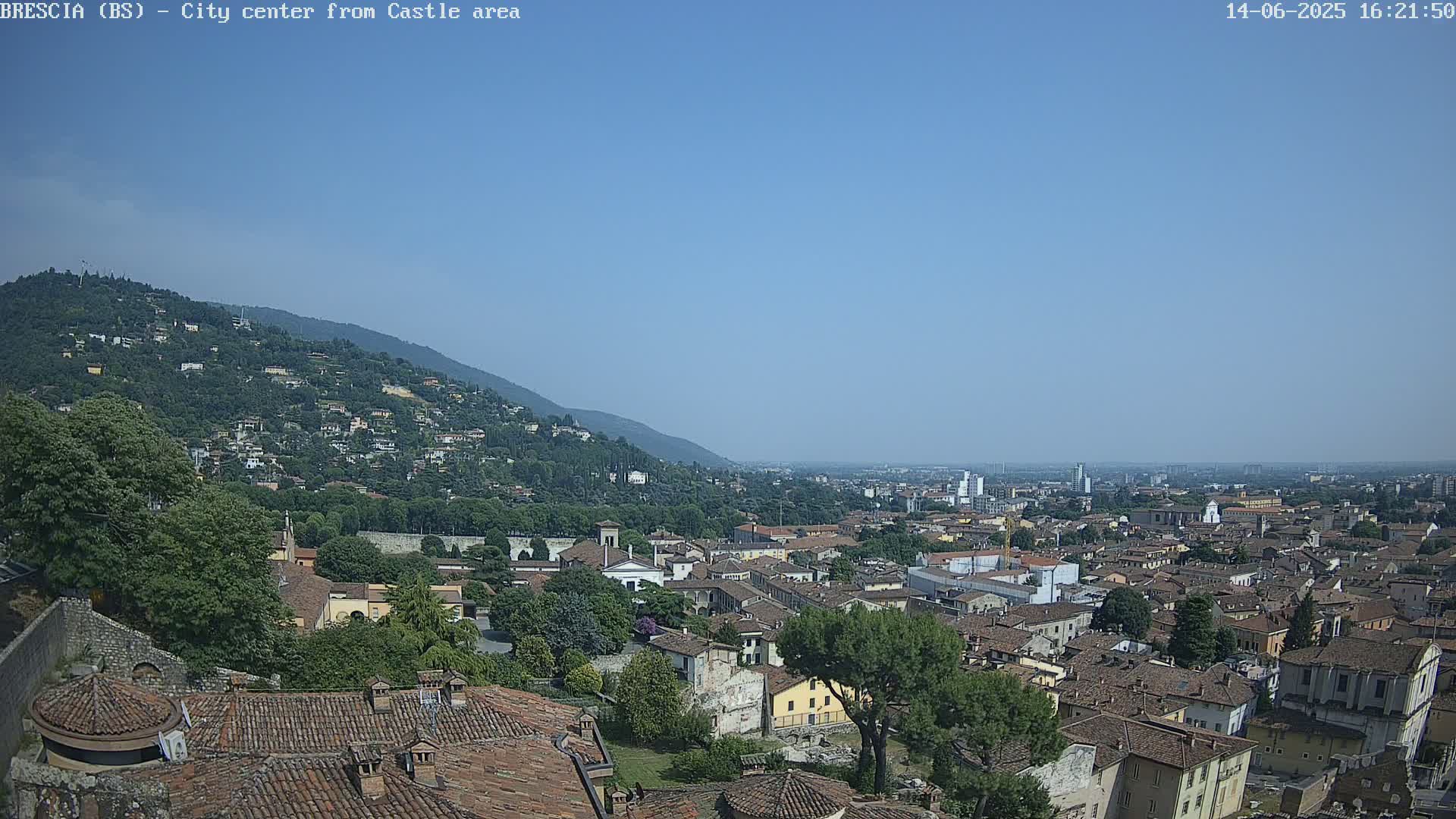 A sprawling city with red-tiled roofs nestled at the base of a verdant hill under a clear blue sky.