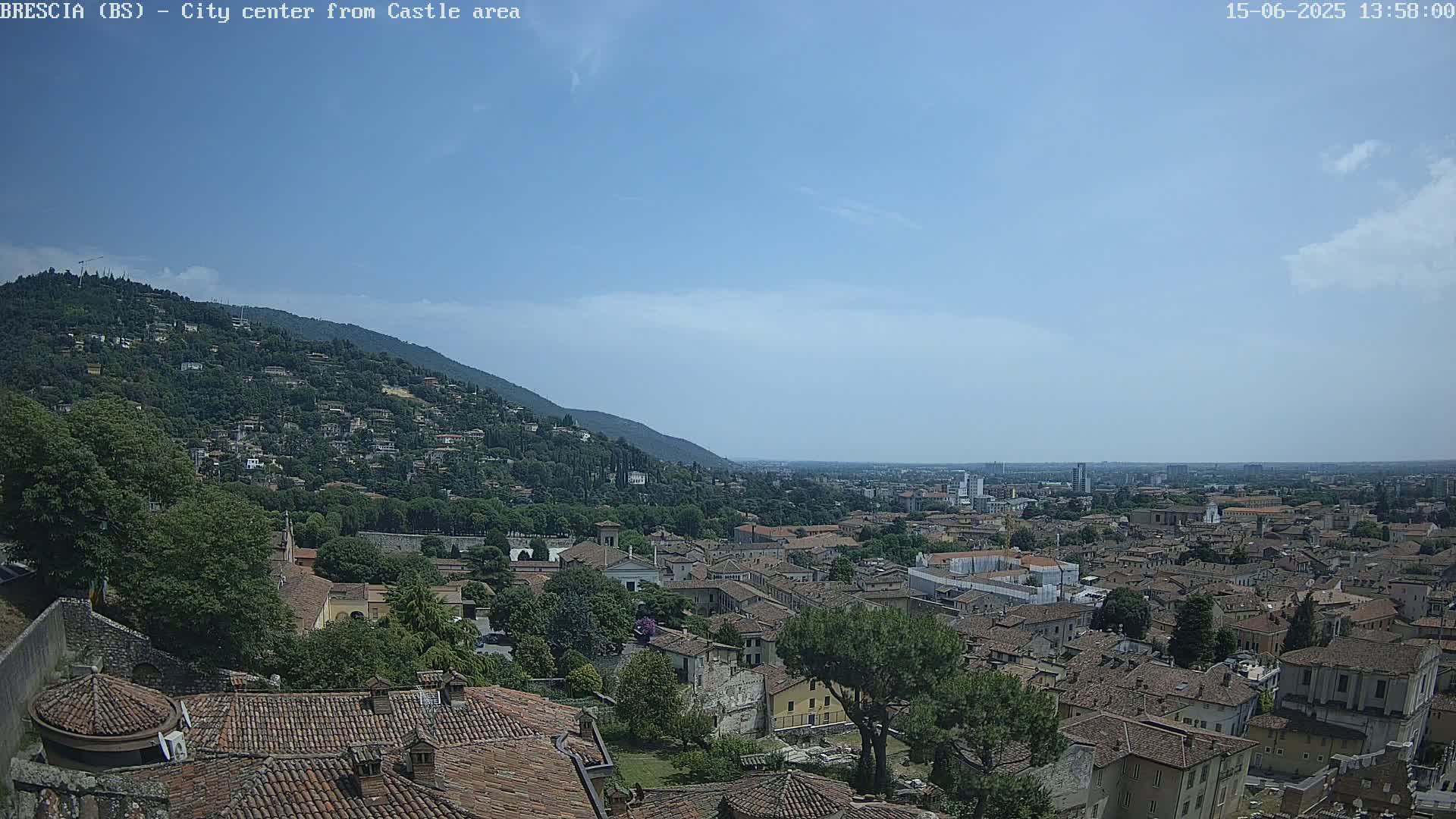 A sprawling city with terracotta rooftops nestles beneath a verdant hill under a mostly clear, bright blue sky.