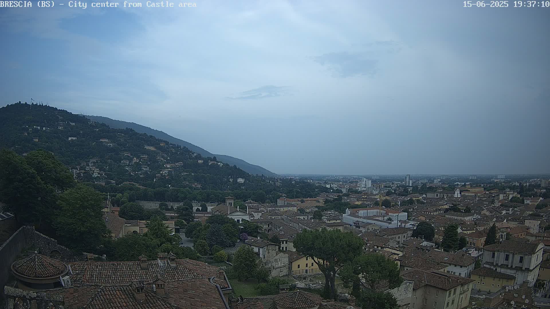 A sprawling city with red-tiled roofs nestled against a verdant hillside under an overcast sky.