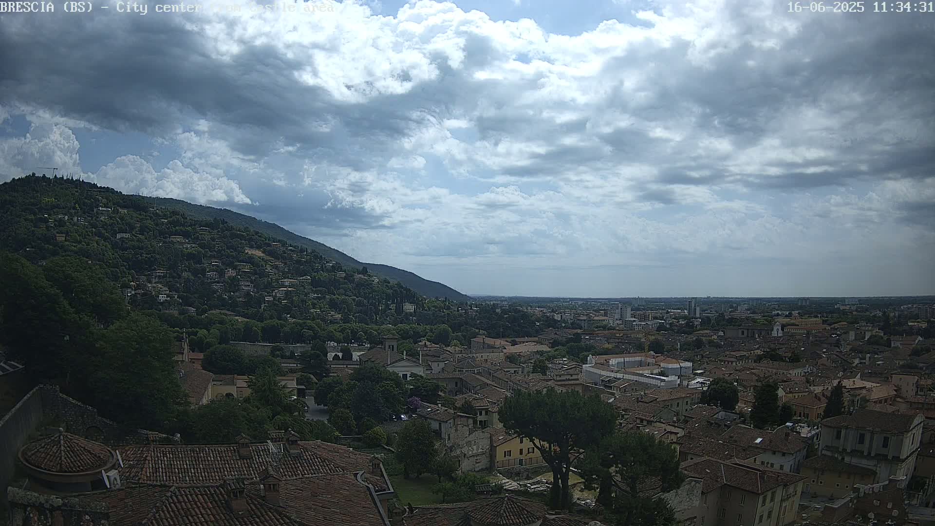 A partly cloudy sky overlooks a city nestled at the base of a green hill, with the distant cityscape stretching to the horizon.