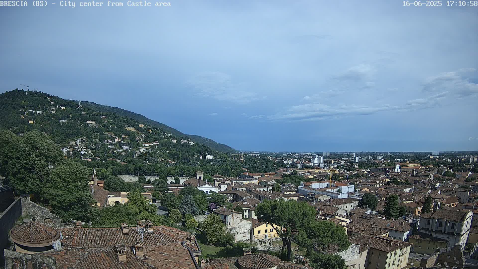 A sprawling city with red-tiled roofs nestled against a green hillside under a mostly clear, light blue sky.