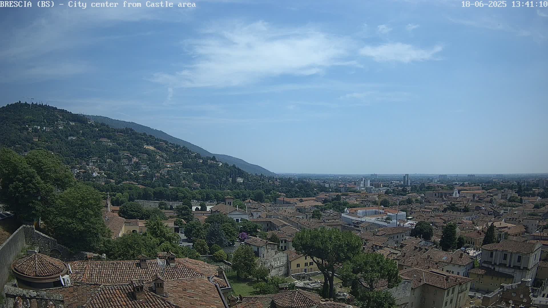 A sprawling city with red-tiled roofs nestled against a green hill under a mostly clear, blue sky.