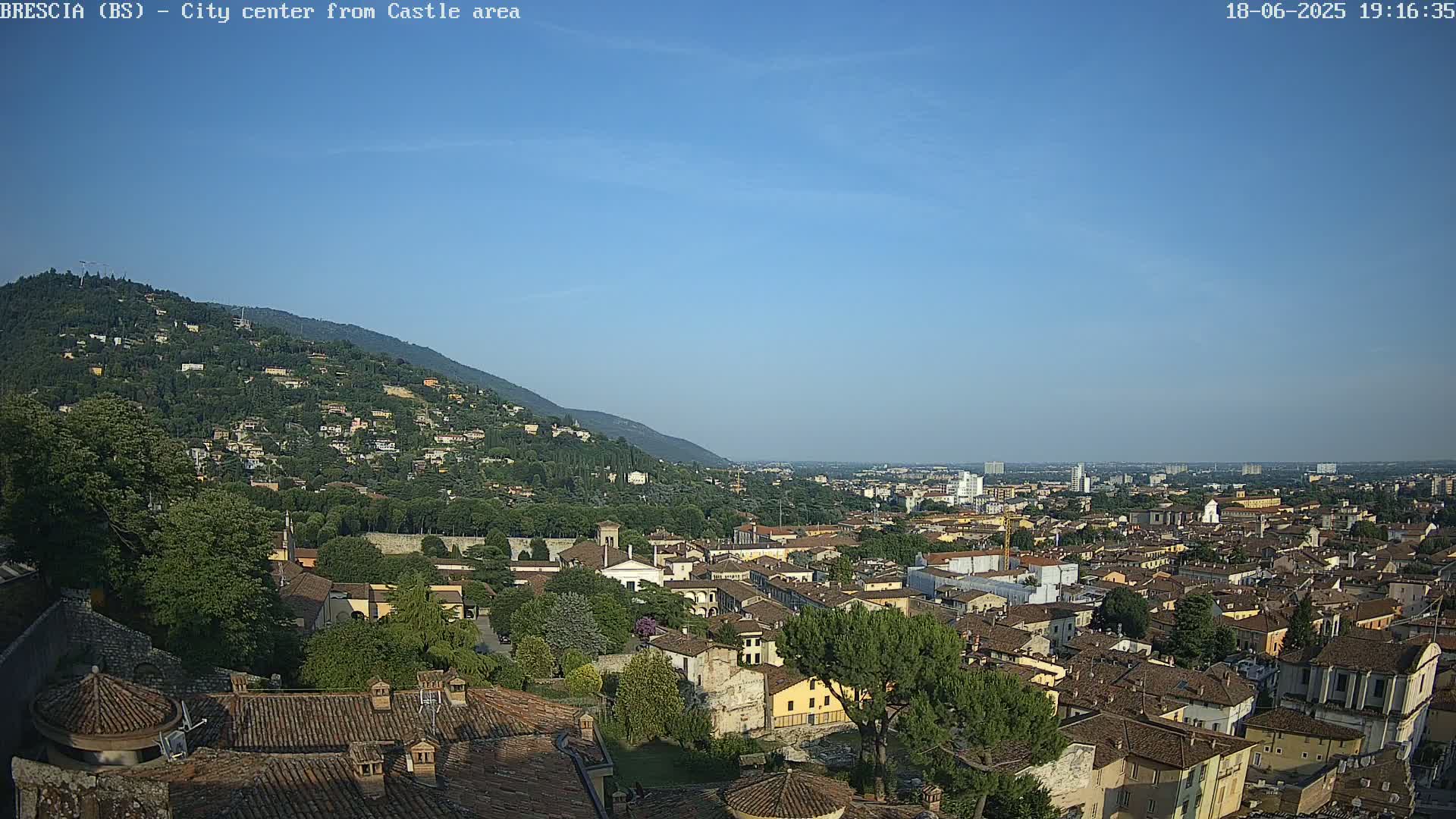 A sprawling city with terracotta rooftops nestles against a verdant hillside under a clear blue sky.