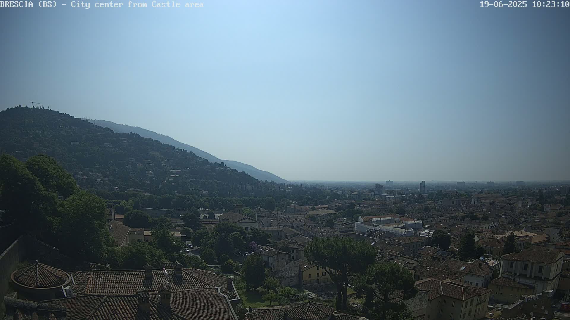 A hazy, sunny day reveals a sprawling city nestled at the foot of a mountain, with terracotta rooftops dominating the foreground.