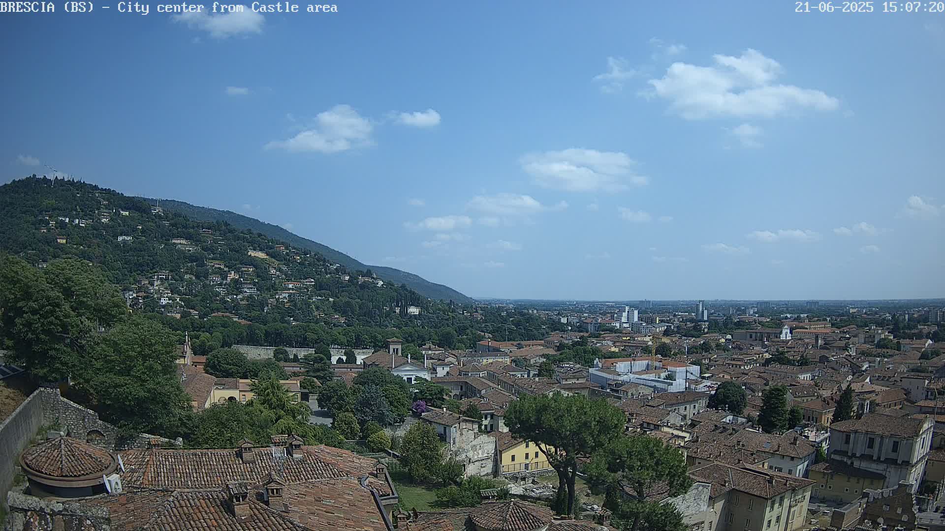 A sprawling city with terracotta rooftops nestles beneath a hill covered in lush greenery under a mostly clear blue sky with a few scattered clouds.