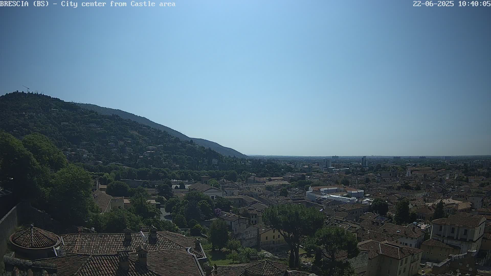 A sprawling city with red-tiled roofs nestles beneath a verdant hill on a clear, sunny day.