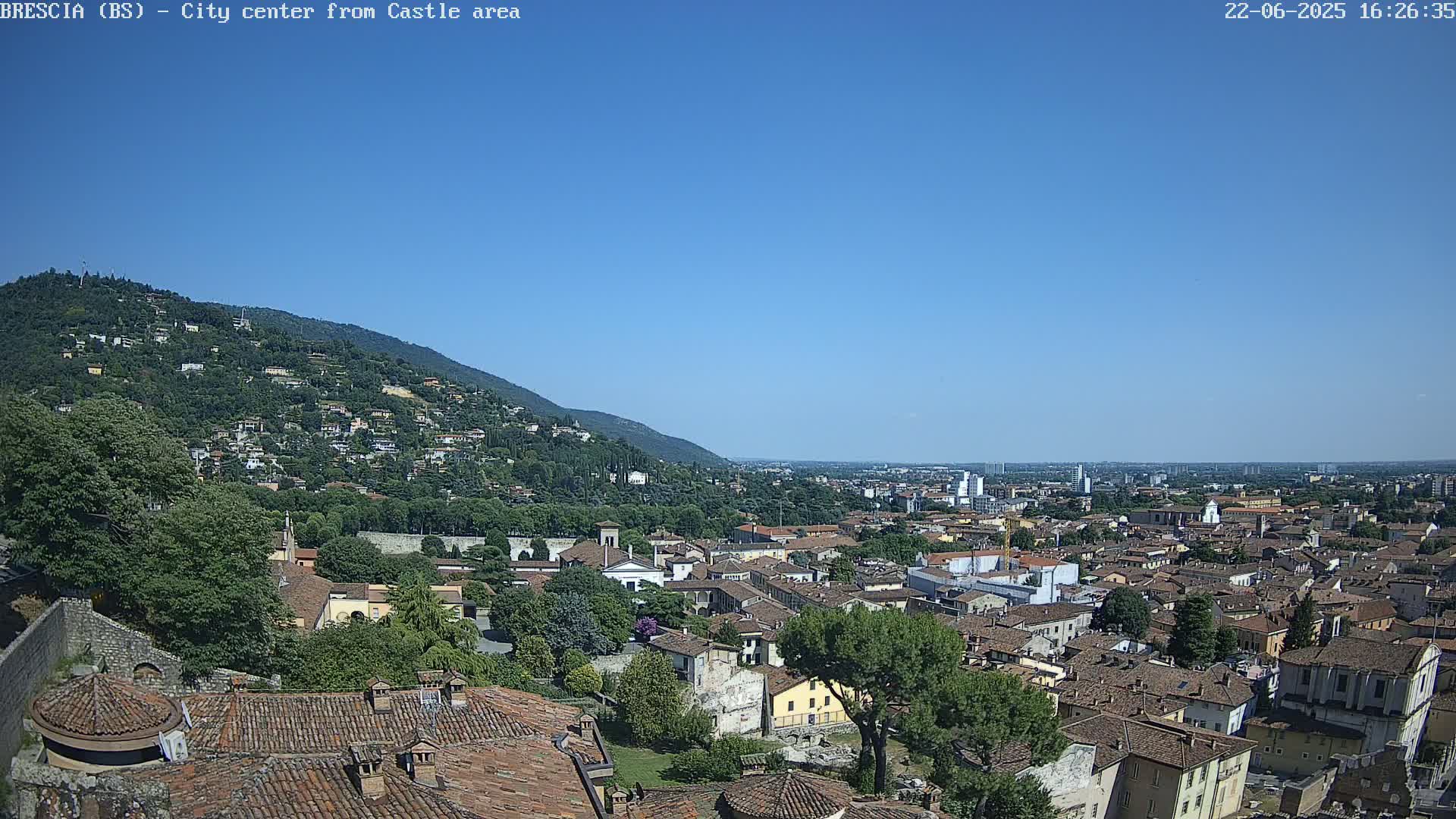 A sprawling city with red-tiled roofs nestled against a verdant hillside under a clear blue sky.