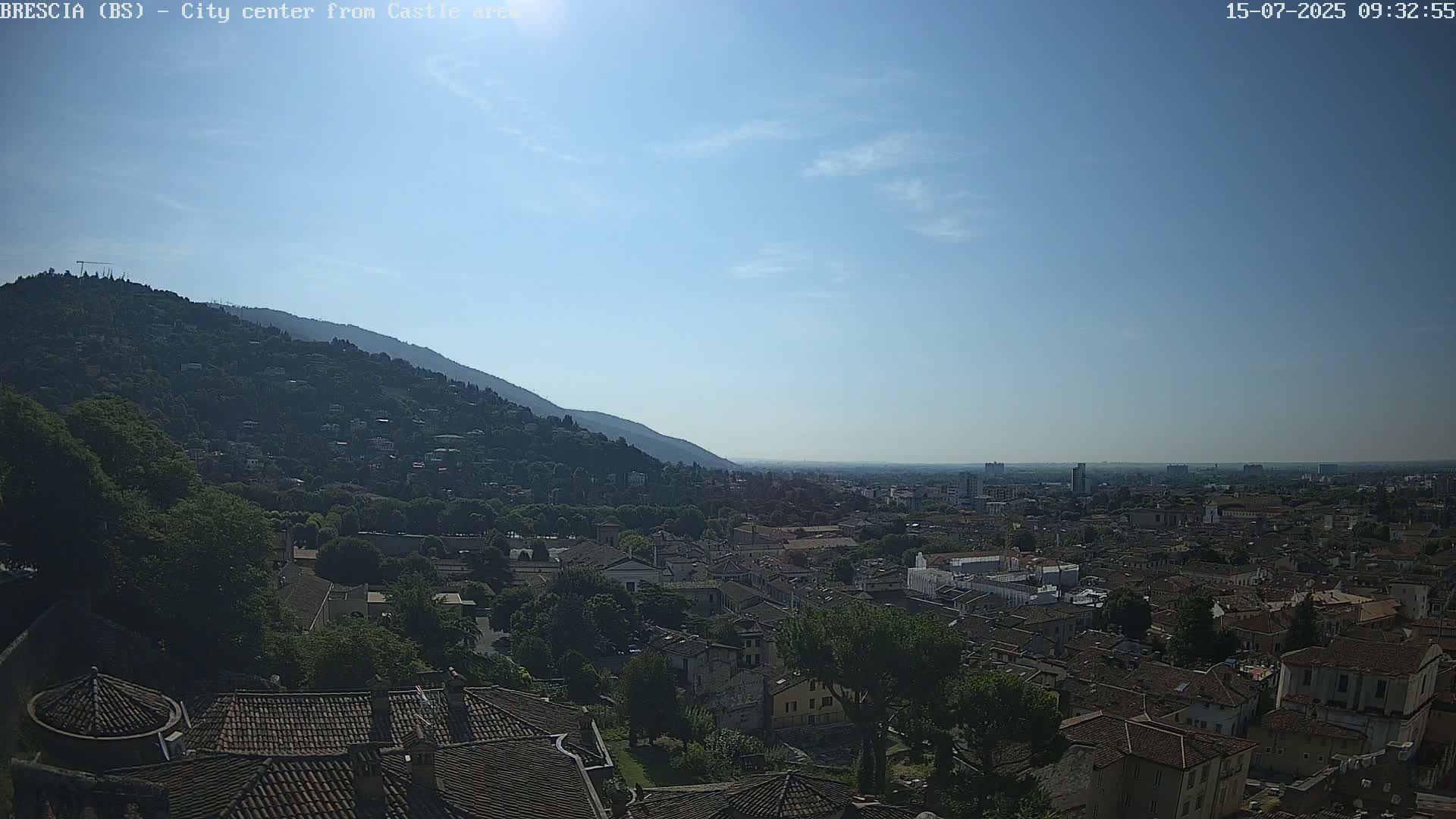 A sprawling city with terracotta roofs nestles at the base of a verdant hill under a clear, sunny sky.