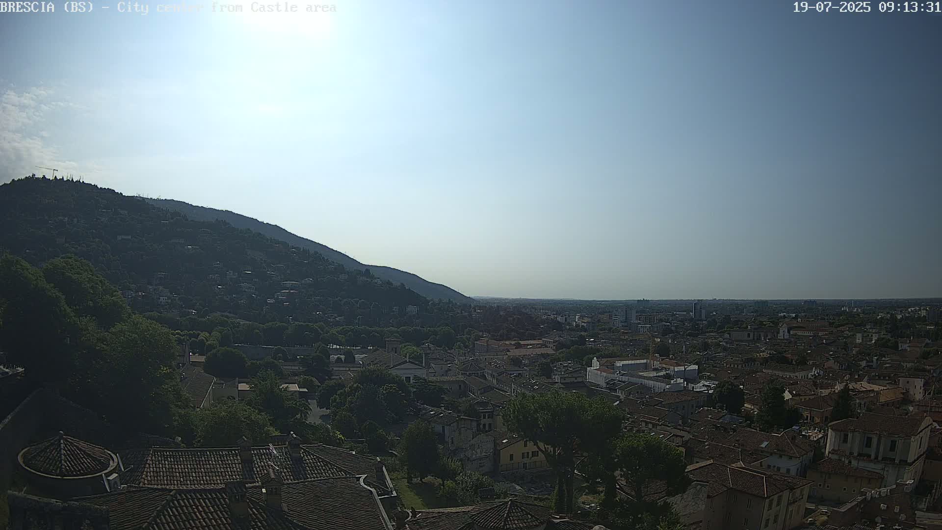 A sprawling city landscape, nestled beneath a hill and under a clear, bright sky, is viewed from an elevated vantage point.