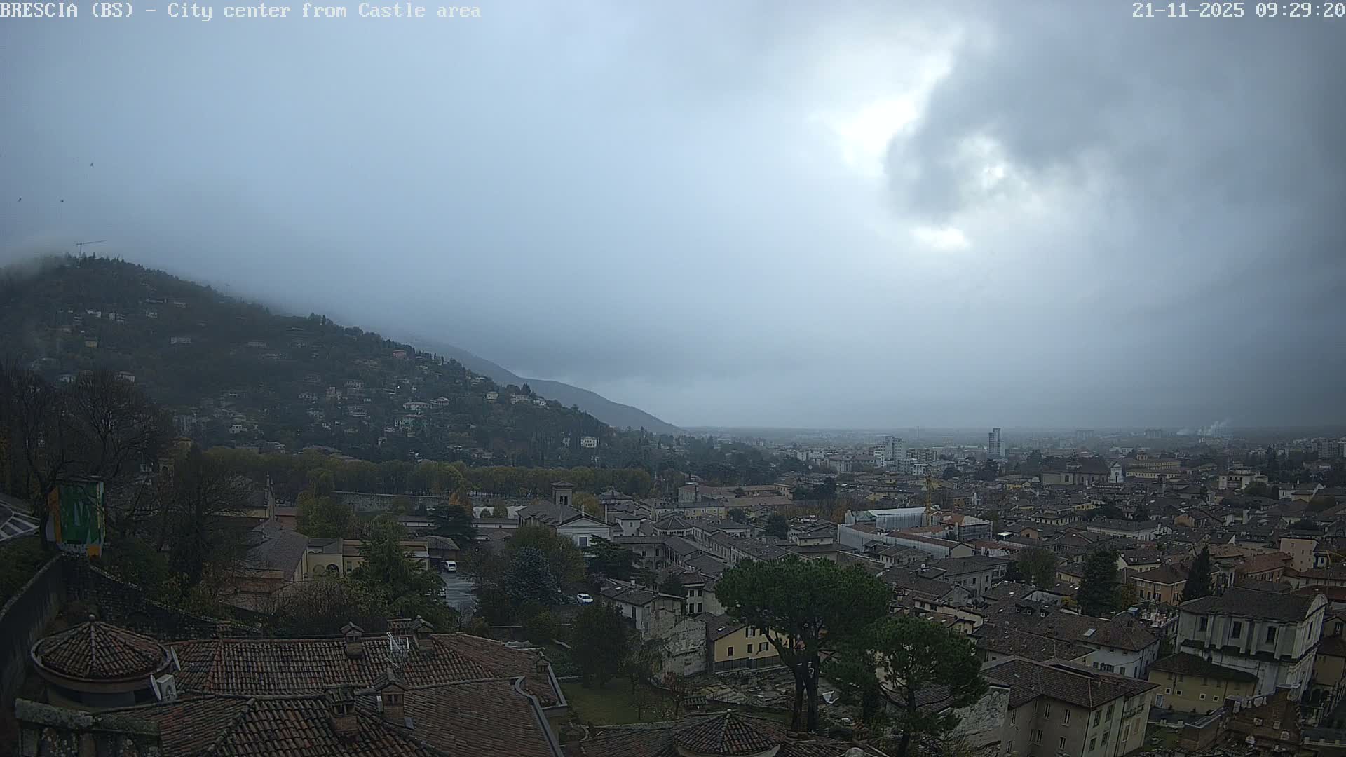 An expansive view of a European city, featuring dense housing on a tree-covered mountain and sprawling across a valley into the misty distance, is captured under a dull, overcast sky with significant haze.