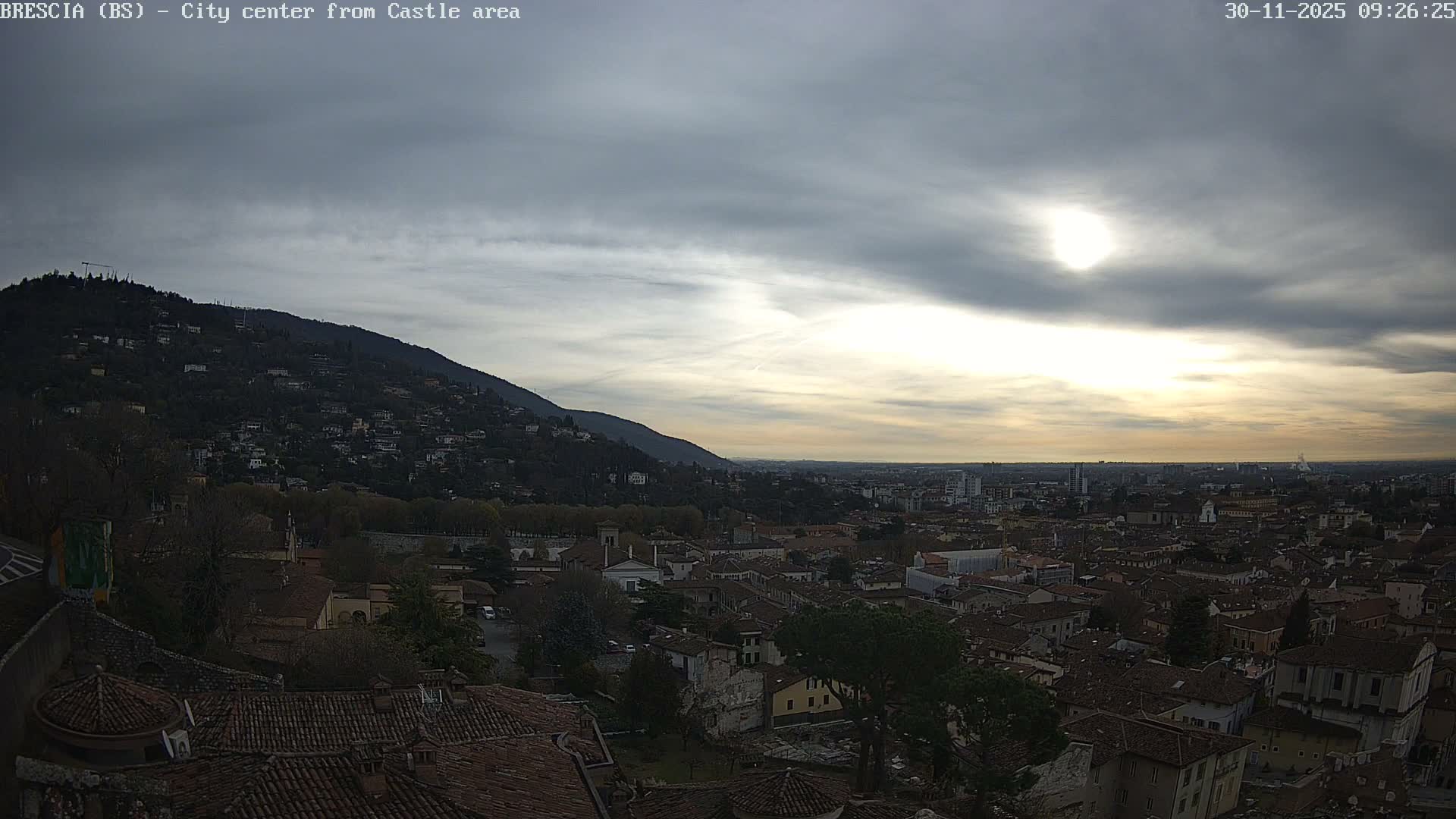 An elevated view reveals a sprawling city of terracotta-tiled rooftops and tree-lined streets set against a large, verdant mountain on the left, all under a mostly cloudy sky with the sun brightly diffused through the clouds.