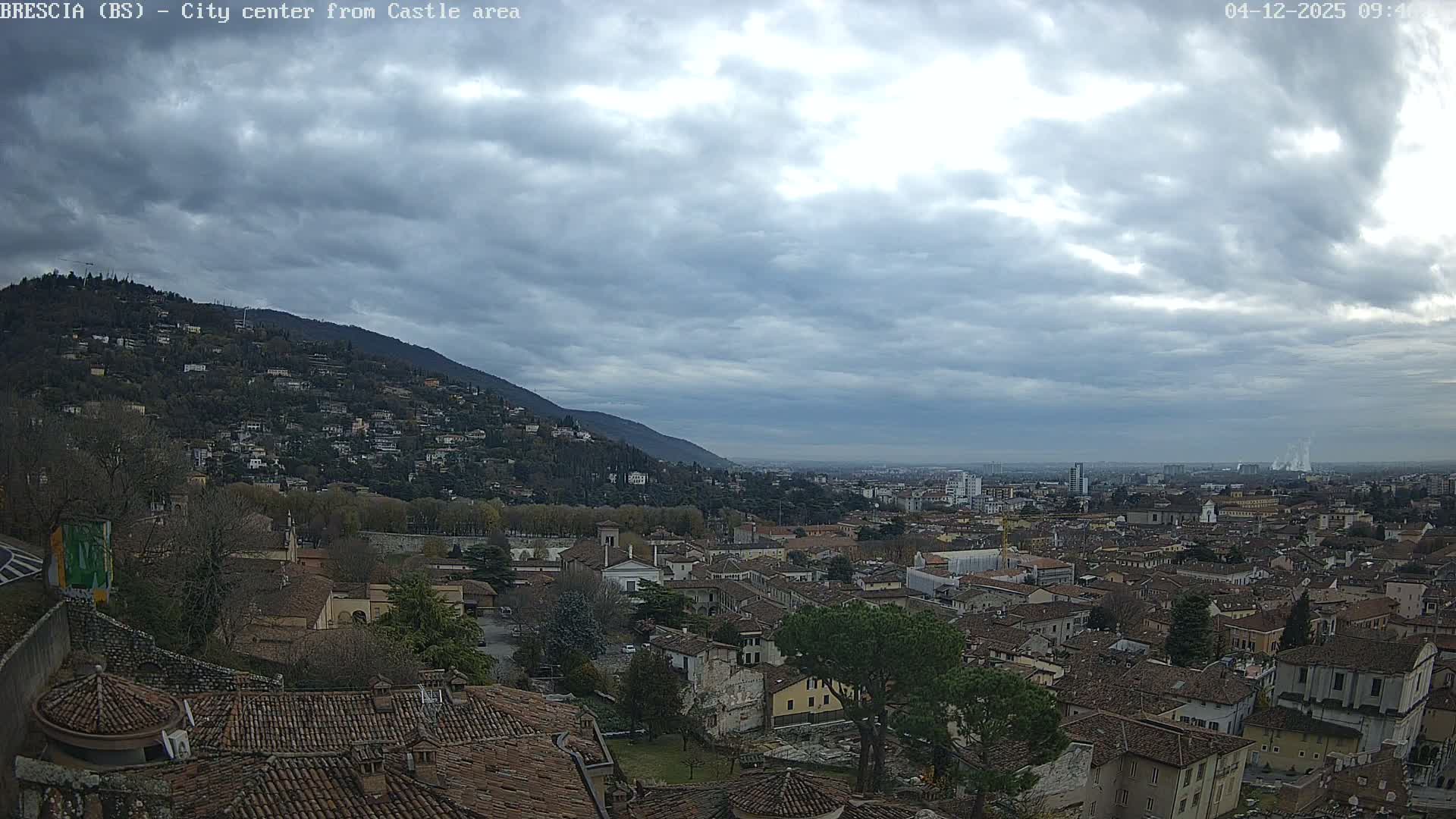 An expansive view of a sprawling city with numerous buildings and terracotta roofs unfolds beneath a heavy, overcast sky, flanked by a tree-covered mountain on the left and industrial plumes rising in the far distance.