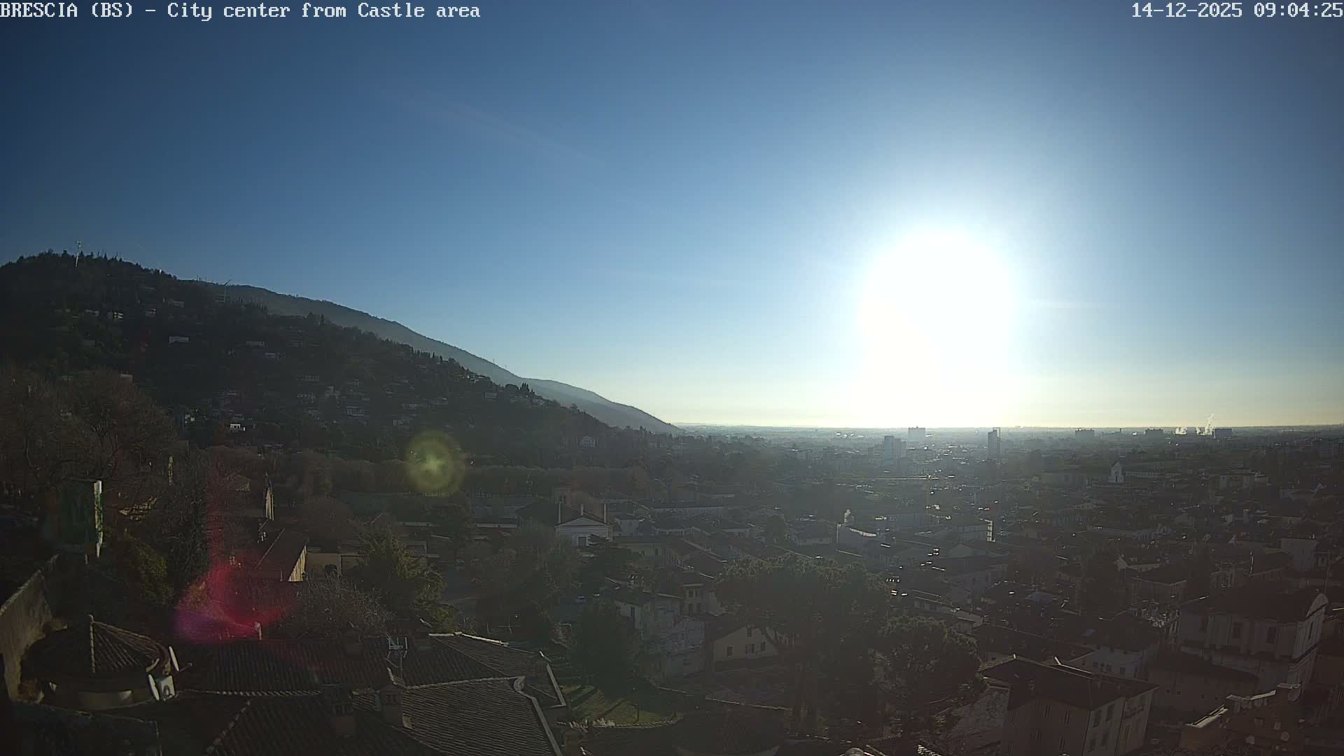 An expansive view reveals a city sprawling at the foot of a tree-covered mountain under a clear blue sky, with a brilliant sun low on the right casting a strong glare and highlighting a hazy distant horizon.