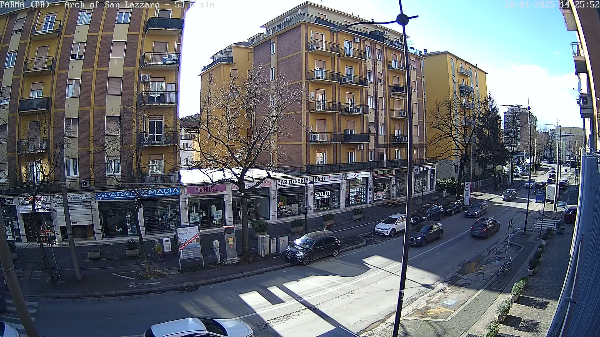 A sunny day on a city street shows several cars parked and driving past multi-story buildings with shops on the ground floor.