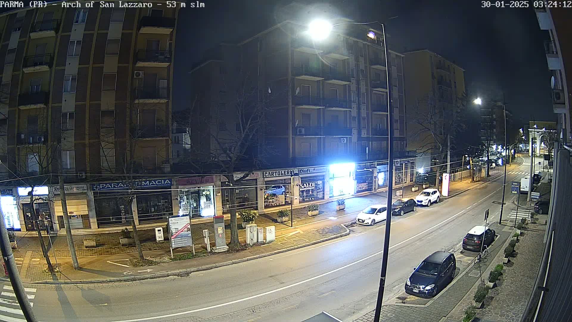 A nighttime street scene shows several parked cars along a multi-lane road in front of shops and a multi-story apartment building under bright streetlights.