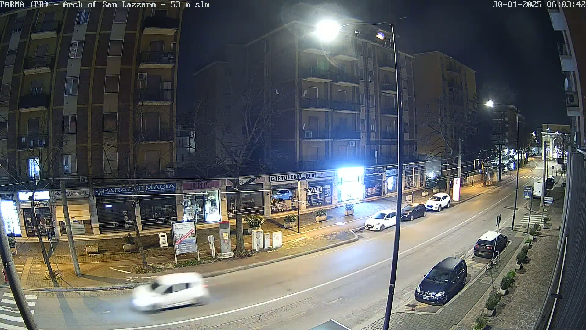 A nighttime street scene shows several parked cars and a few moving vehicles on a road lined with buildings, under bright streetlights.
