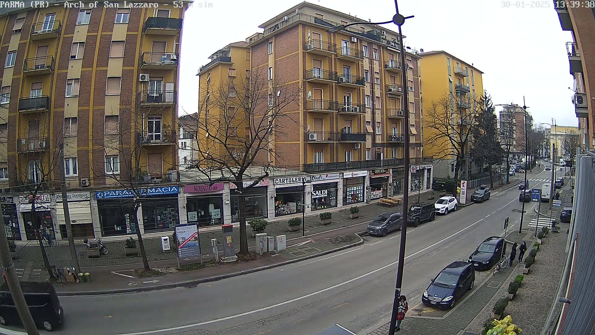 A city street scene on an overcast day shows a multi-story yellow building with shops on the ground floor, cars parked along the street, and bare trees lining the sidewalk.