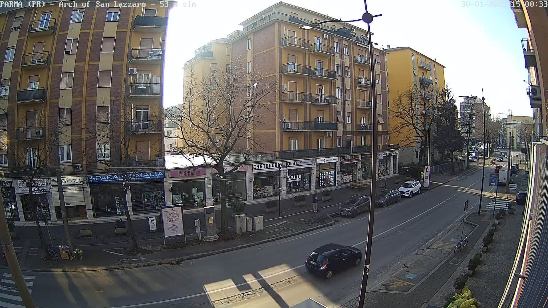 A sunny street scene shows a multi-story brick building with shops on the ground floor, several parked cars, and a few cars driving down the street.