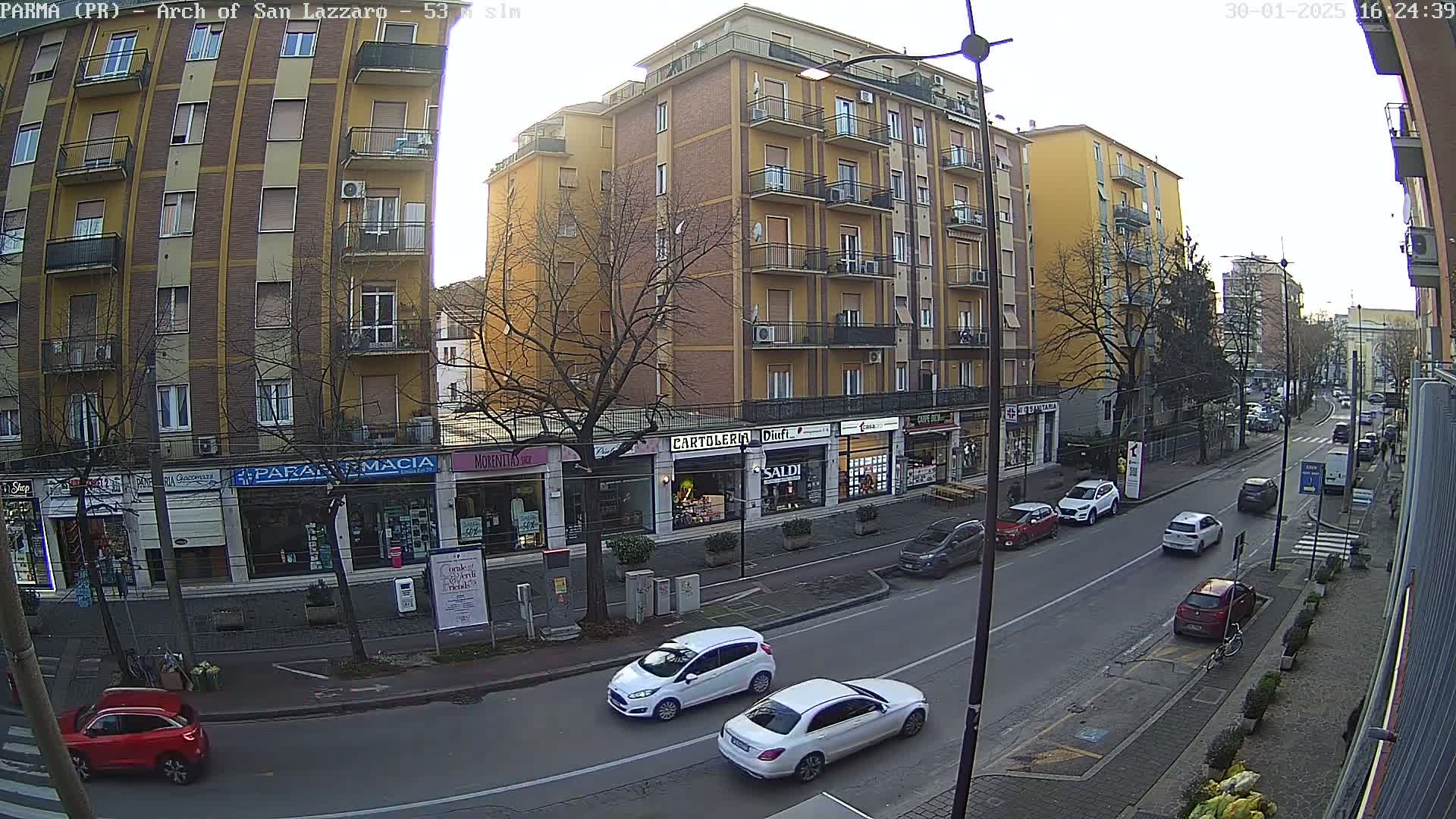 A street scene on a partly sunny day shows several cars parked and driving along a city street lined with multi-story buildings and shops.