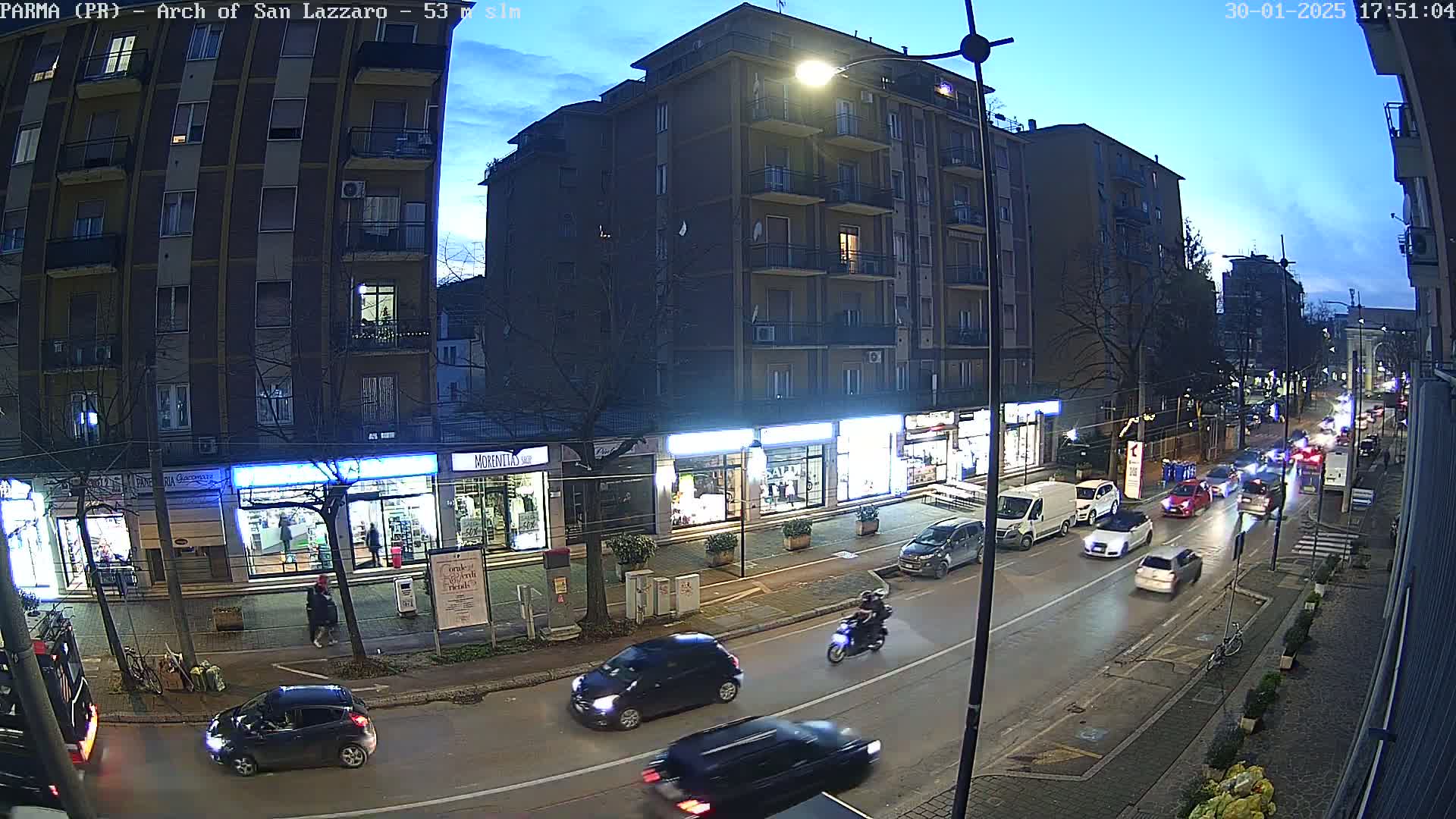 A twilight view of a city street with cars driving on a wet road, passing shops and buildings under a partly cloudy sky.