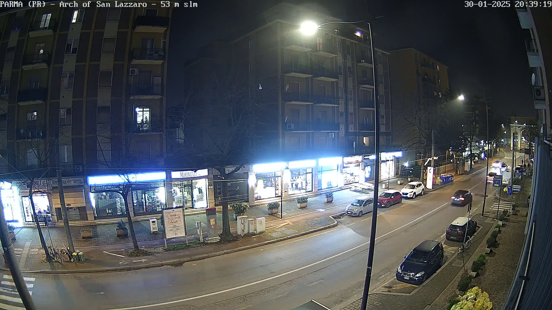 A nighttime street scene shows several parked and moving cars in front of illuminated storefronts, next to a tall apartment building under streetlights.
