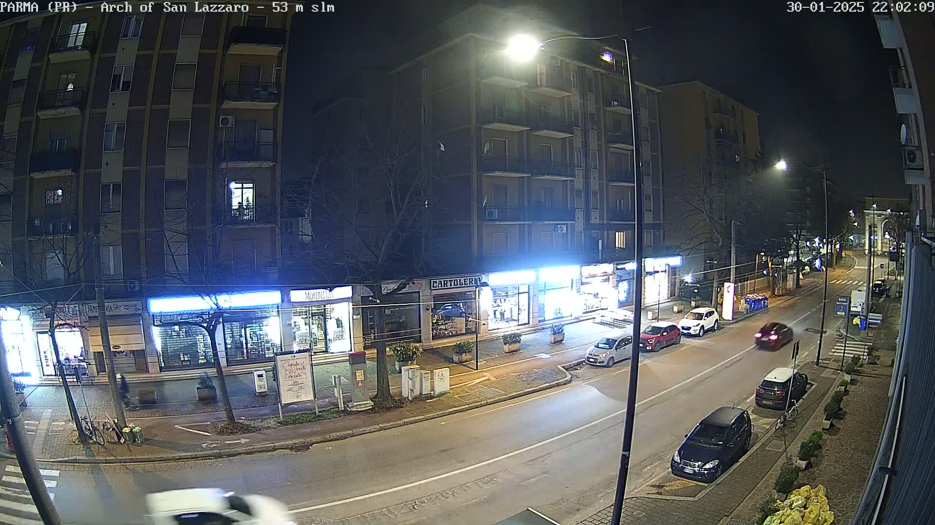 A nighttime street scene shows several parked and moving cars in front of closed shops and a multi-story apartment building under streetlights.