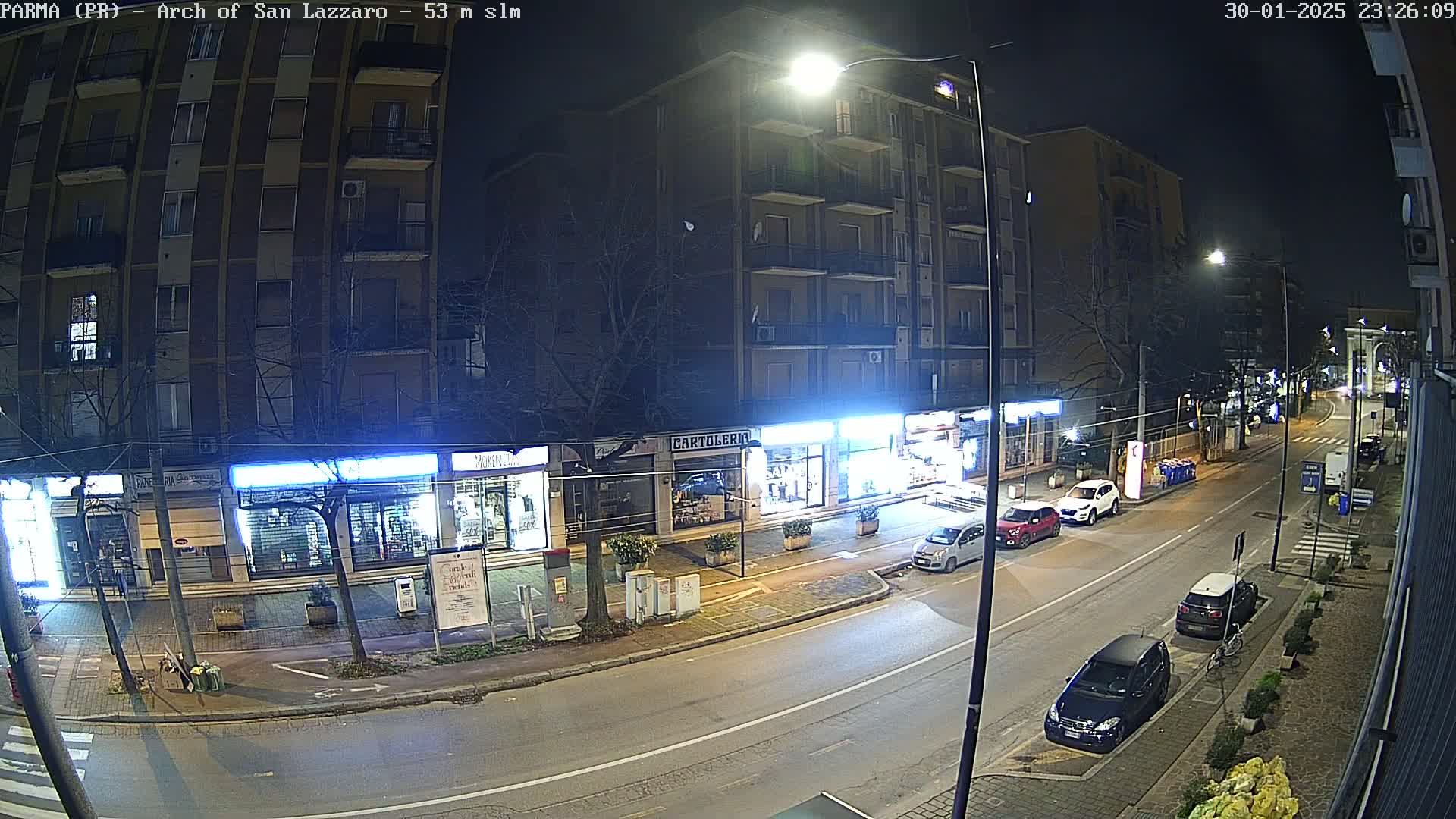 A nighttime street scene shows several cars parked along the side of a road in front of stores and apartment buildings, under streetlights.