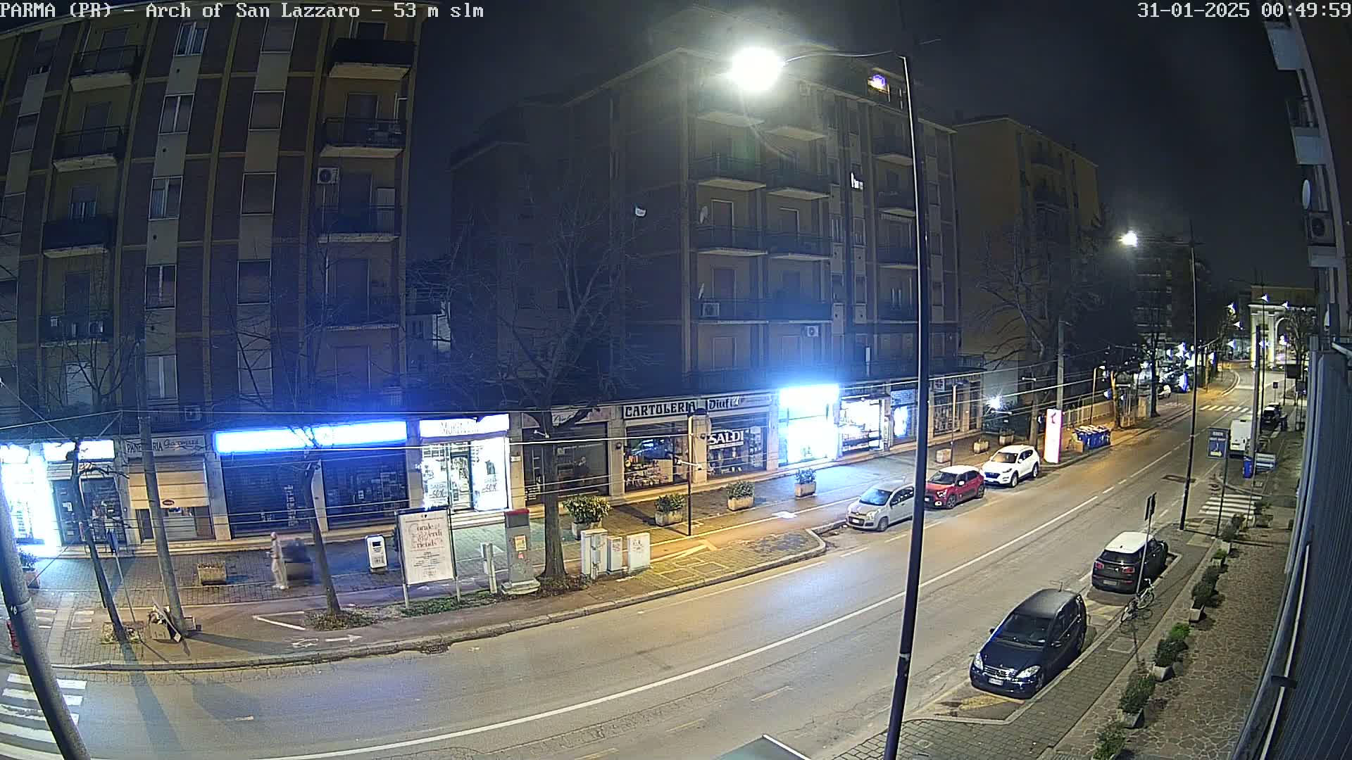 A nighttime street scene shows several cars parked along the curb in front of shops and a multi-story building under streetlights.