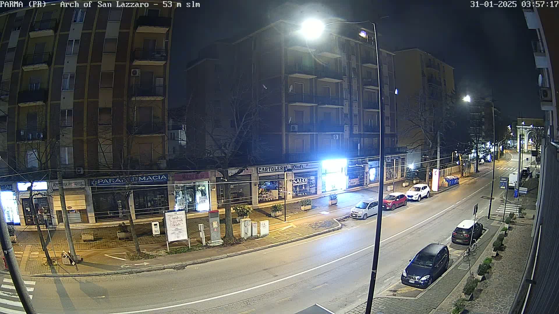 A nighttime street scene shows several parked cars along the side of a road lined with shops and a multi-story building under bright streetlights.