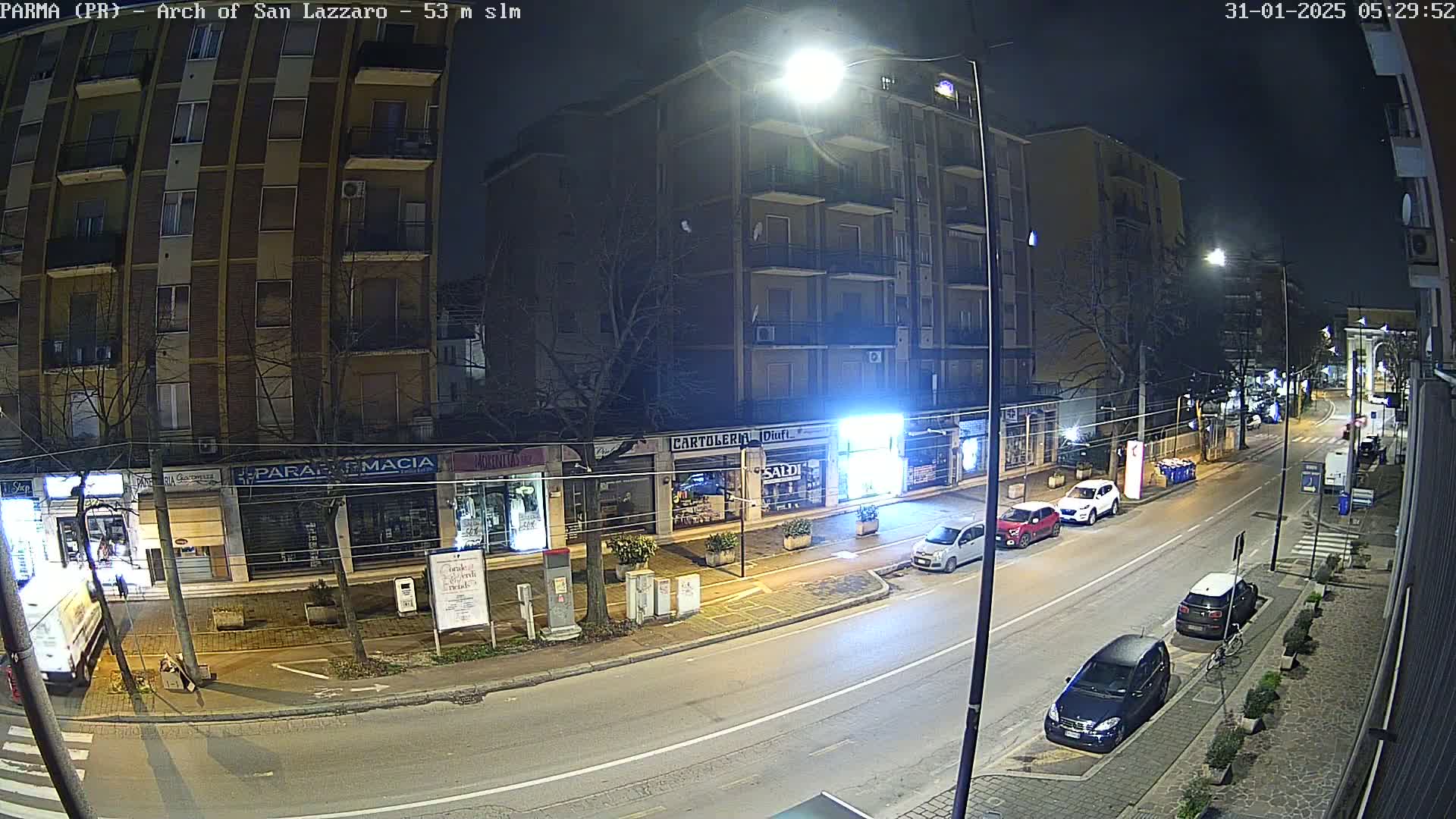 A nighttime street scene shows several parked cars and a few driving cars on a roadway lined with shops and a multi-story building.