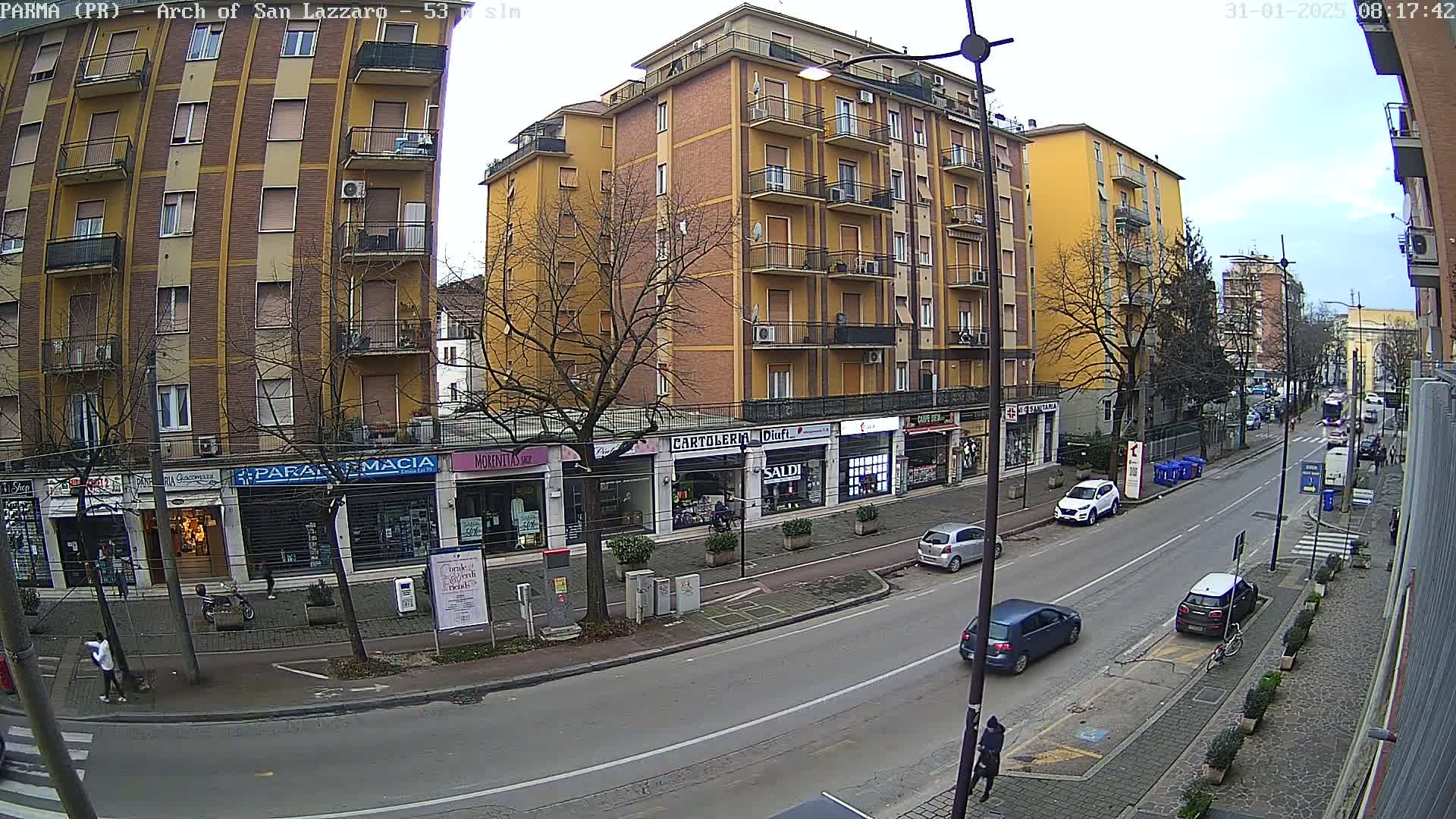 A mostly sunny city street scene shows a multi-story yellow building with shops on the ground floor, several parked cars, and a few pedestrians.