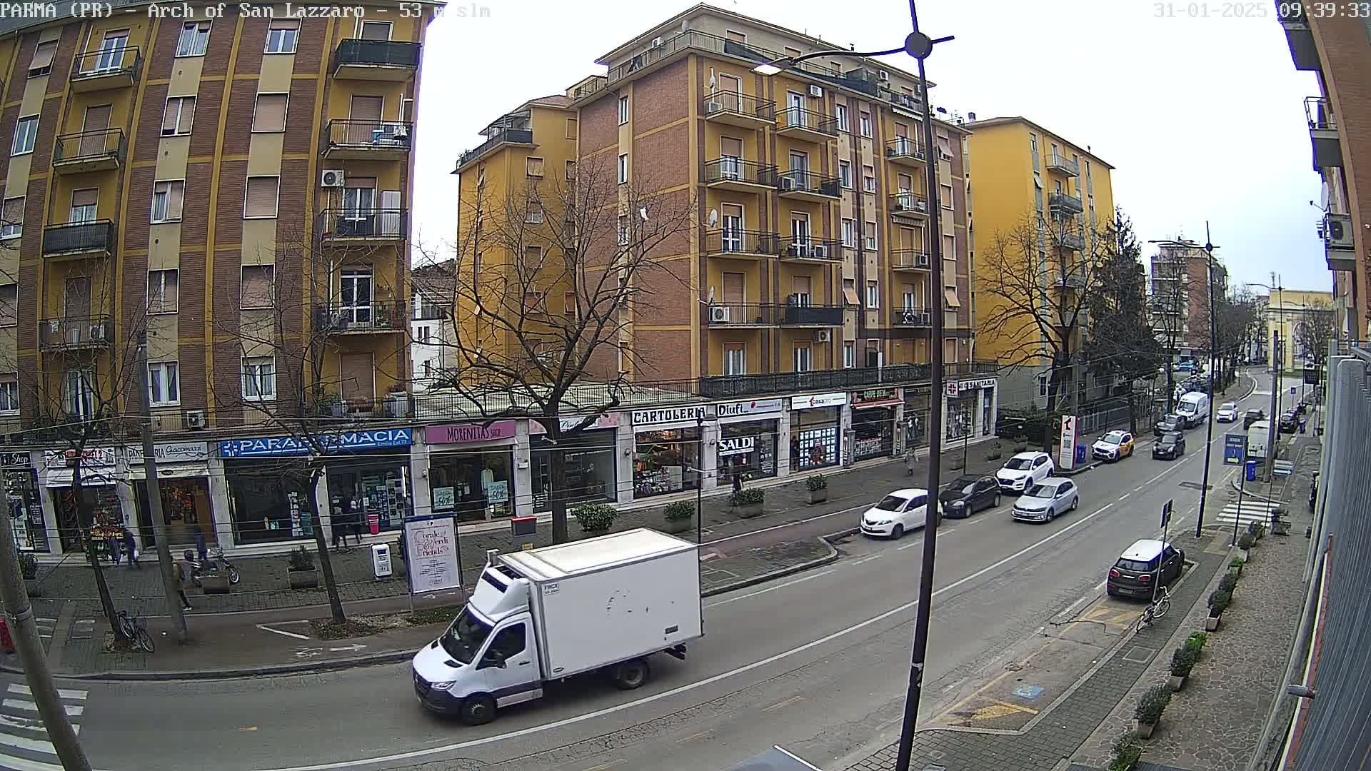 A city street scene on an overcast day shows a row of shops and apartment buildings, with cars parked along the side of the street and a white delivery van in the foreground.