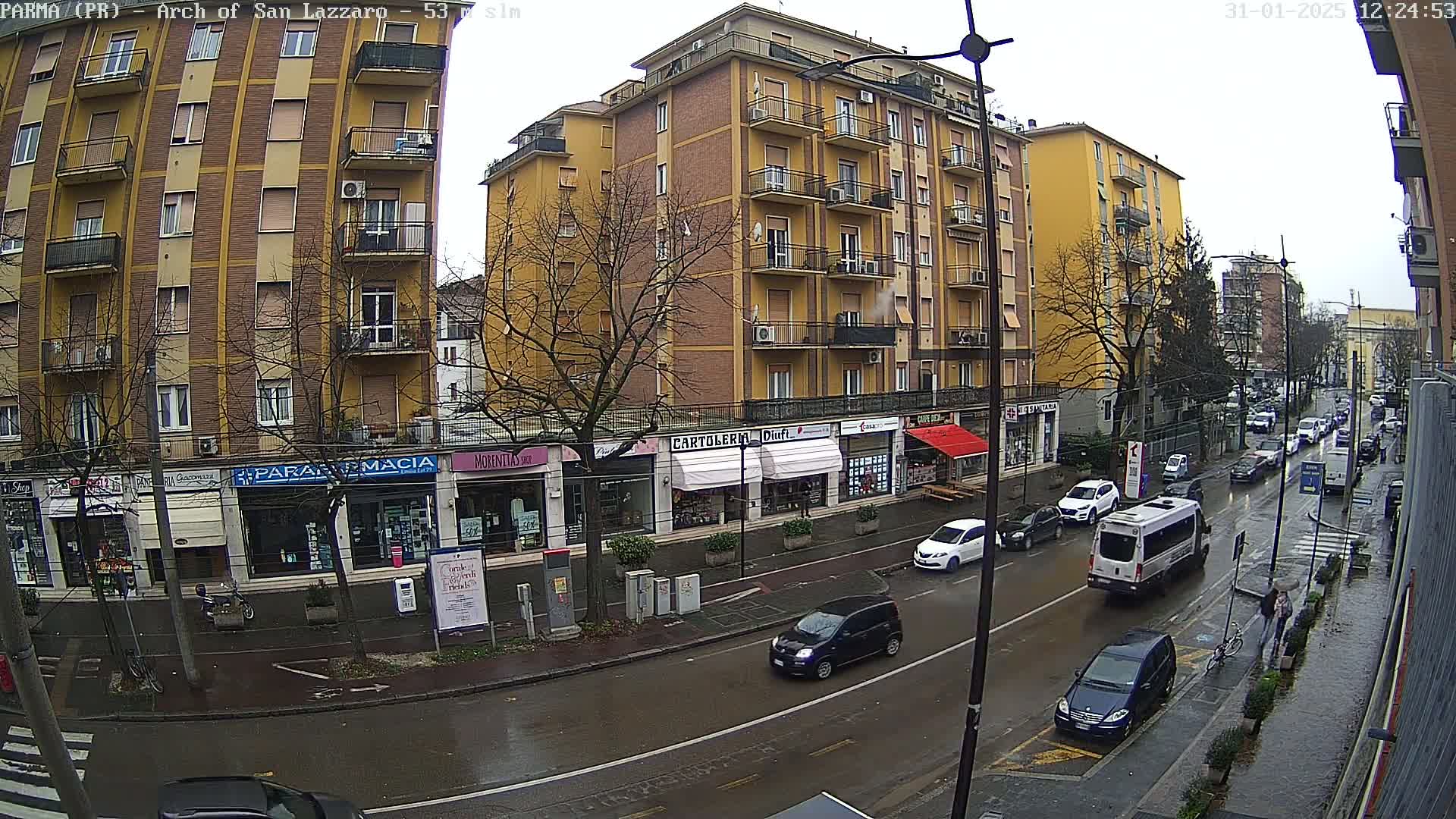 A rain-slicked city street scene shows a multi-story yellow building with shops on the ground floor, several cars driving on the street, and pedestrians walking along the sidewalk.