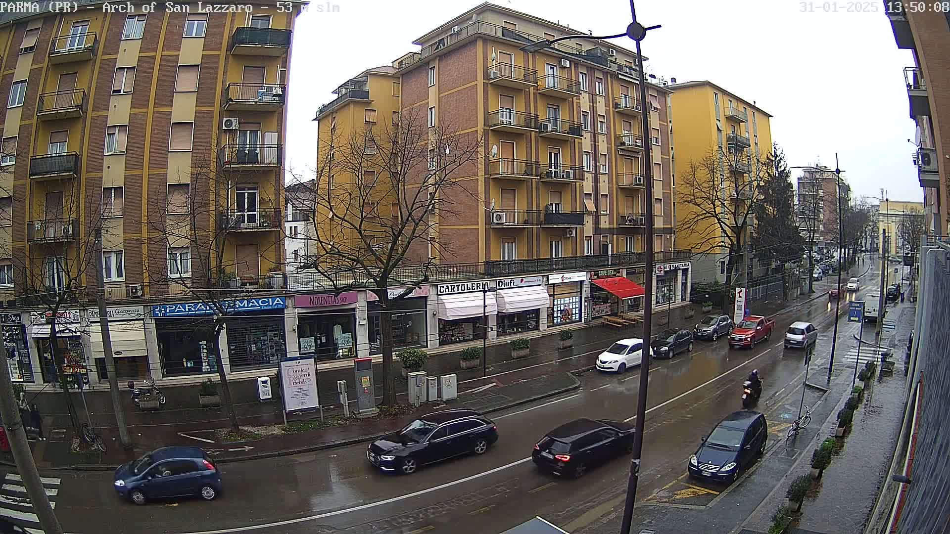 A wet city street scene shows several cars driving past shops and a tall yellow building under an overcast sky.