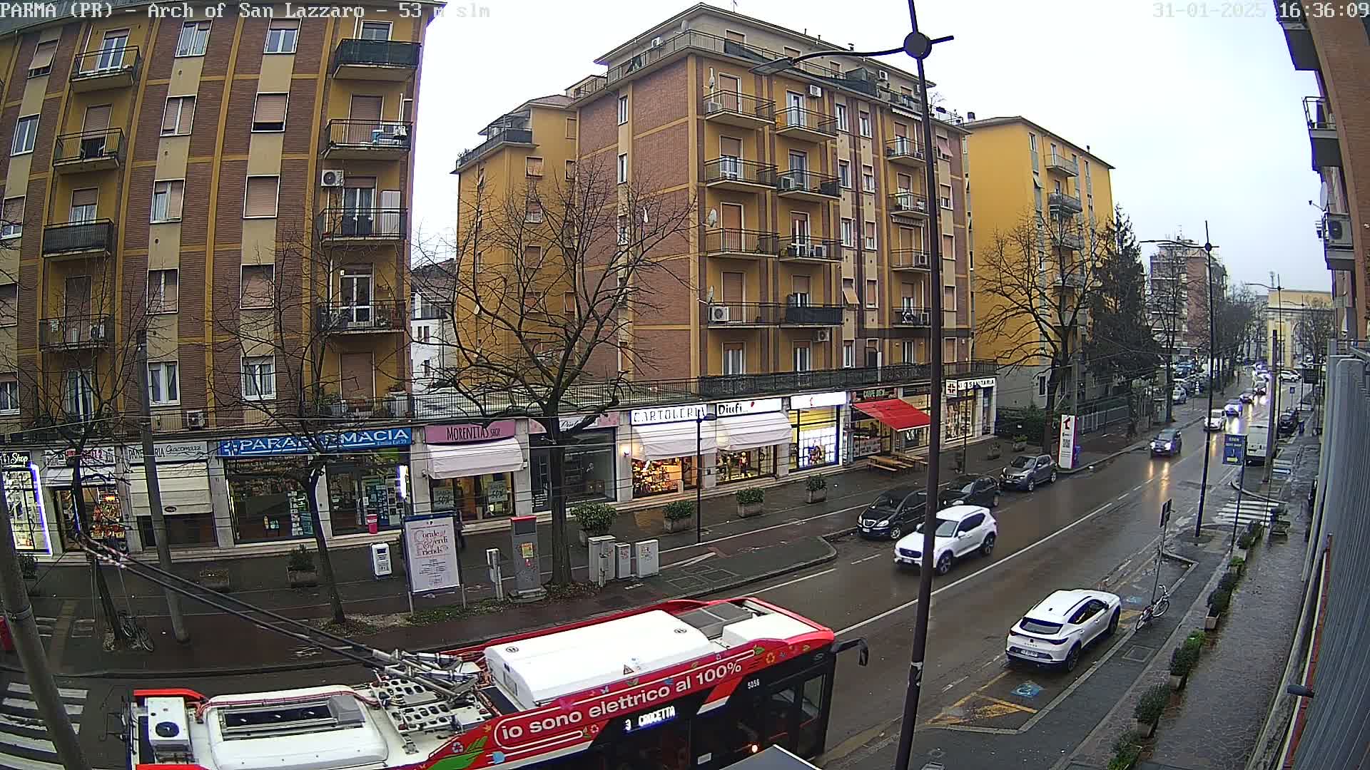 A wet city street scene shows a multi-story building and shops along the street, with cars and a bus, under an overcast sky.