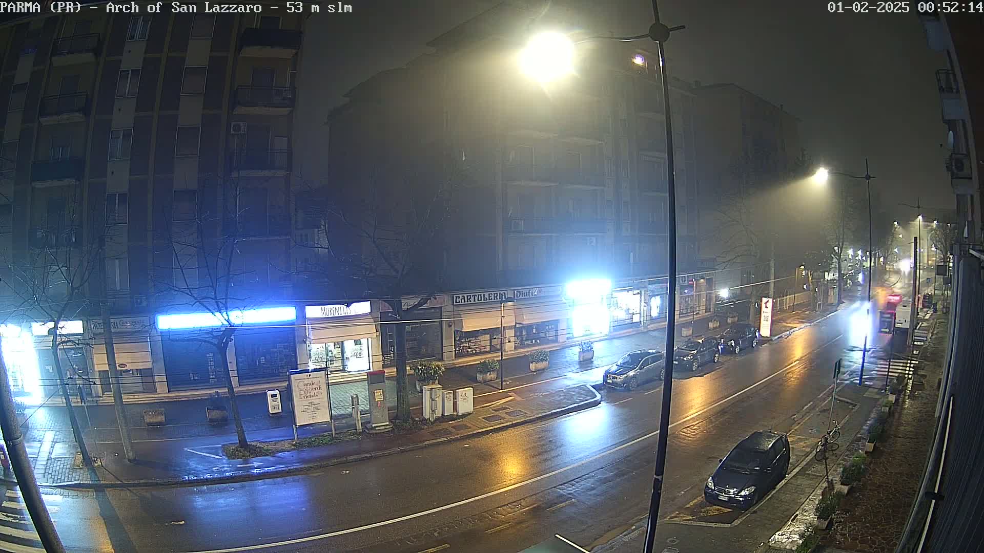 A wet, foggy street at night, showing several buildings with illuminated storefronts and a few cars parked along the side of the road.