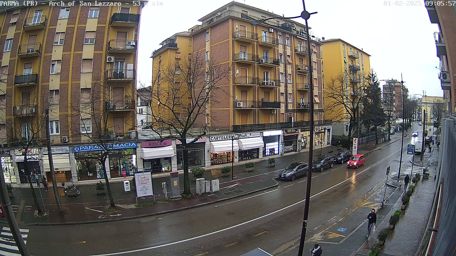 A wet city street scene shows several parked cars and pedestrians on a rainy day, with multi-story buildings and shops lining both sides of the road.